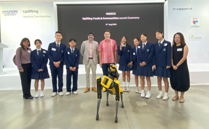 Group of people in school uniforms posing with a yellow Boston Dynamics robot dog at a Hyundai launch ceremony.