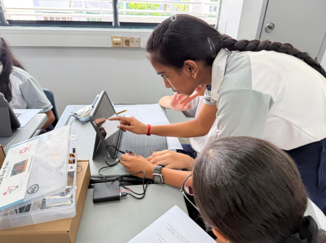 Two students collaborate at a desk, with one pointing at a tablet screen while the other types on a laptop keyboard.