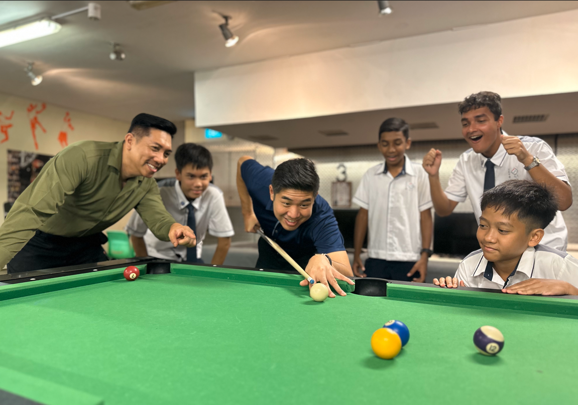 A man in blue leans over a green pool table to take a shot while five others watch and cheer around him.