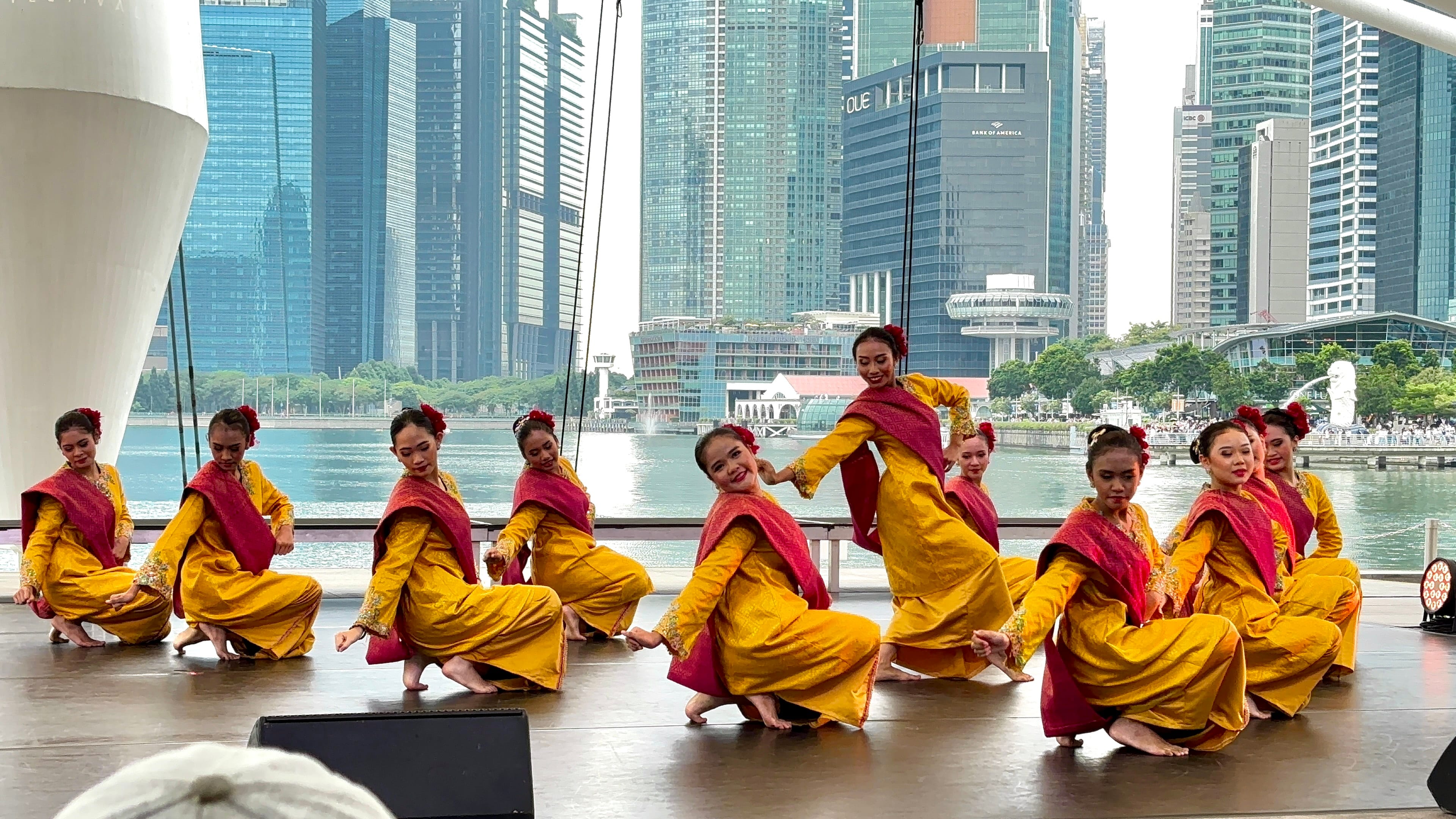 Women in yellow and red traditional costumes perform a cultural dance on a stage with Singapore's skyline behind them.