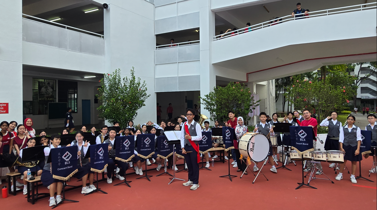 Students in uniforms from BLS Concert Band pose outdoors in a school courtyard with instruments and music stands.
