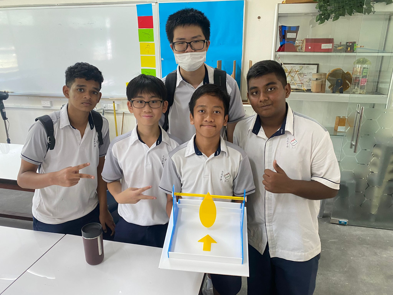 Five male students in school uniforms pose together holding a blue and yellow model project in a classroom.