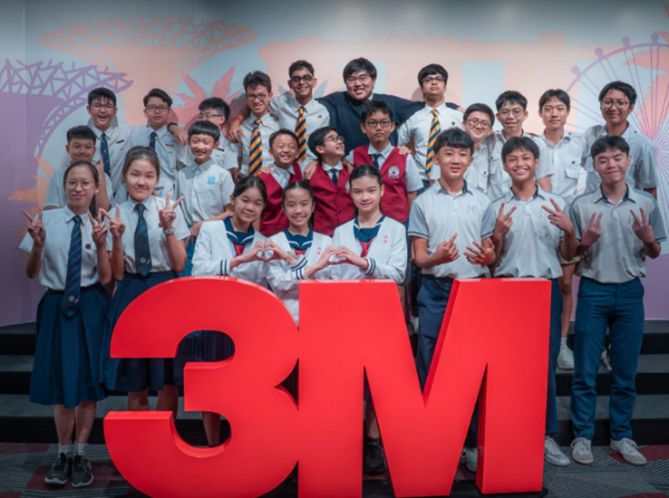 A group of school students in uniforms pose behind a large red 3M logo sign, making hand gestures.