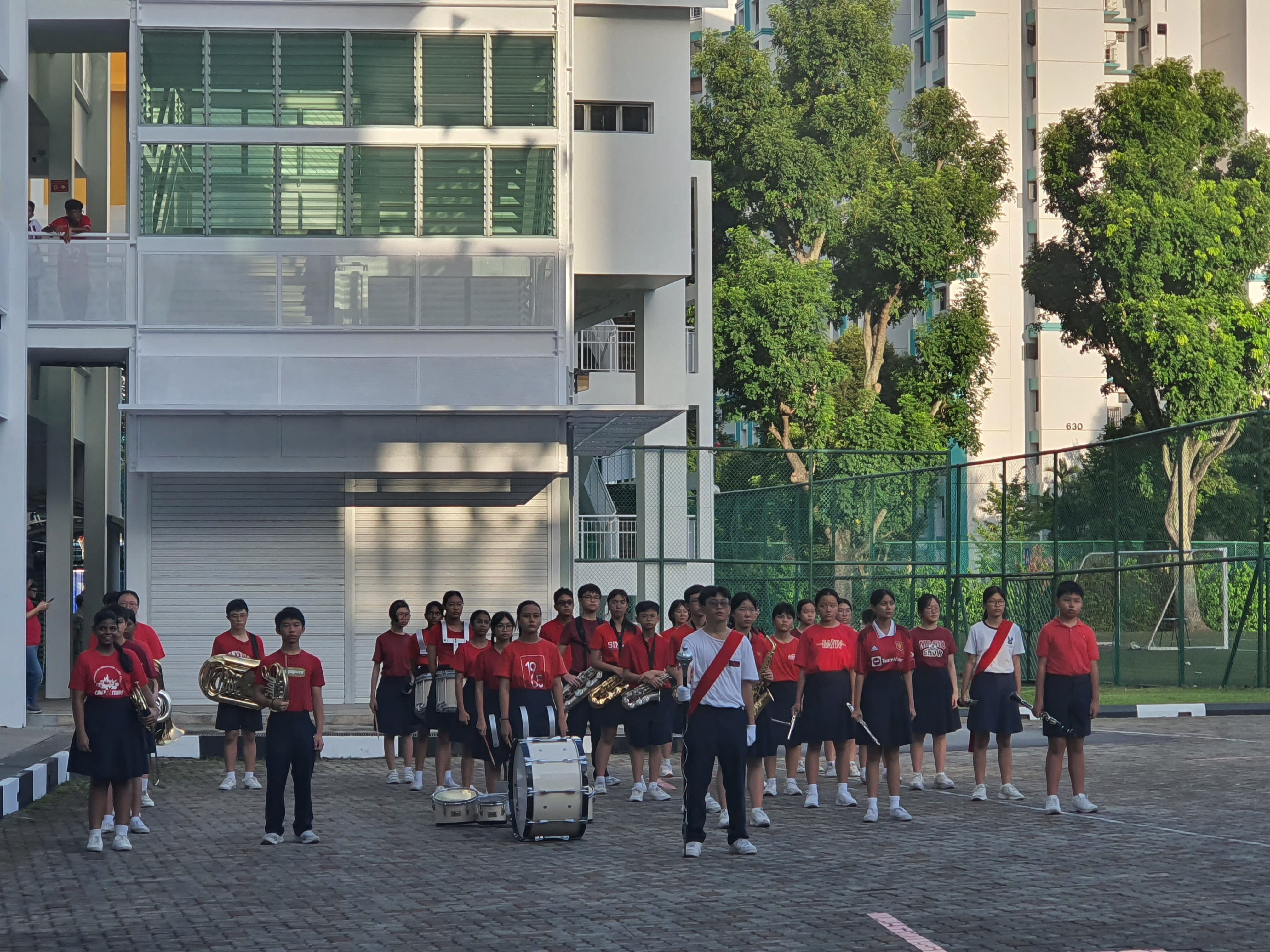 Students in red uniforms stand in formation outdoors with brass and drum instruments beside a white building.