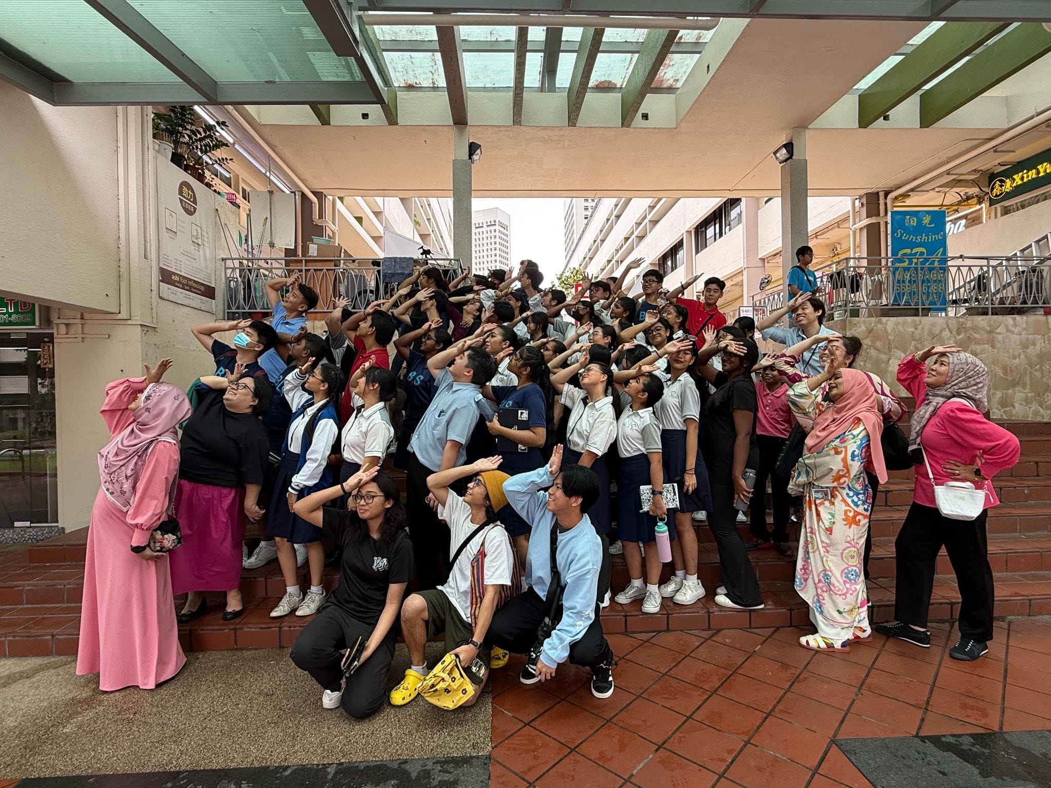A large group of students and adults pose outdoors, shielding their eyes and looking upward together.