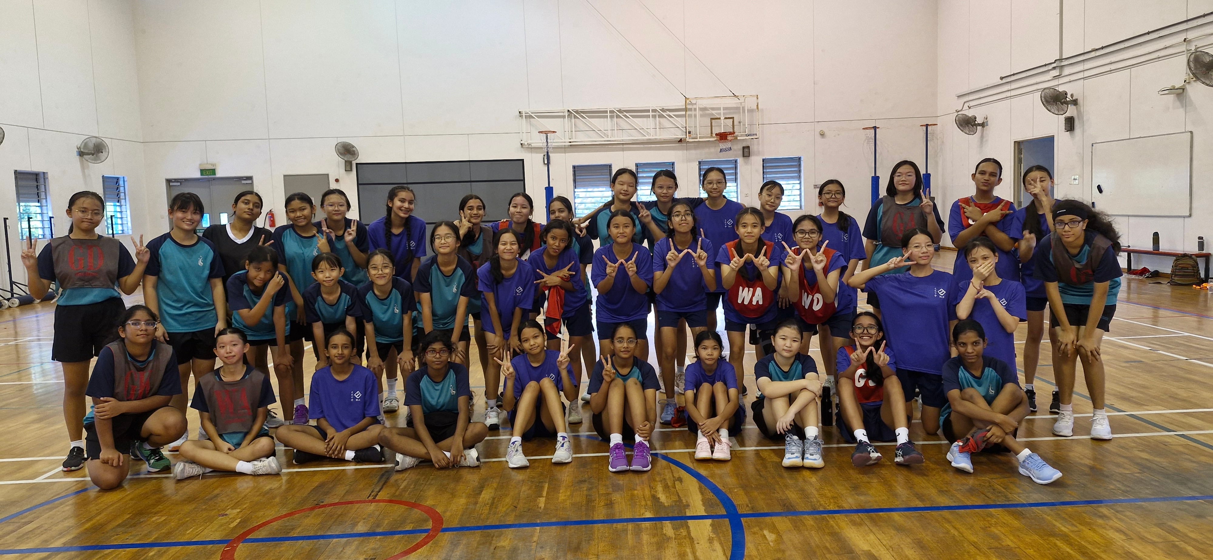 A large group of girls in blue and teal sports uniforms posing with peace signs inside an indoor gymnasium.