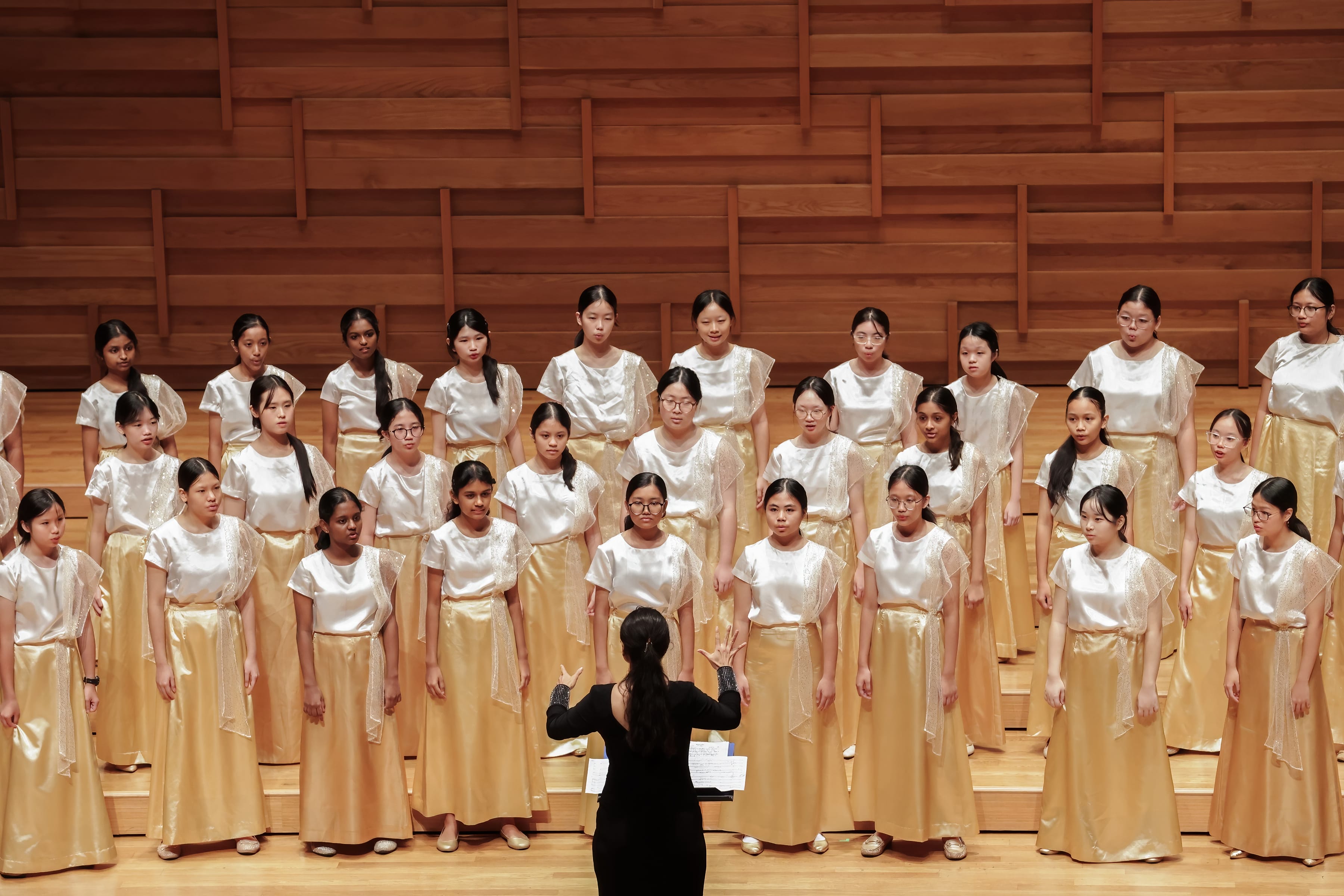 A conductor in black leads a large girls choir in white tops and gold skirts on a wooden stage.