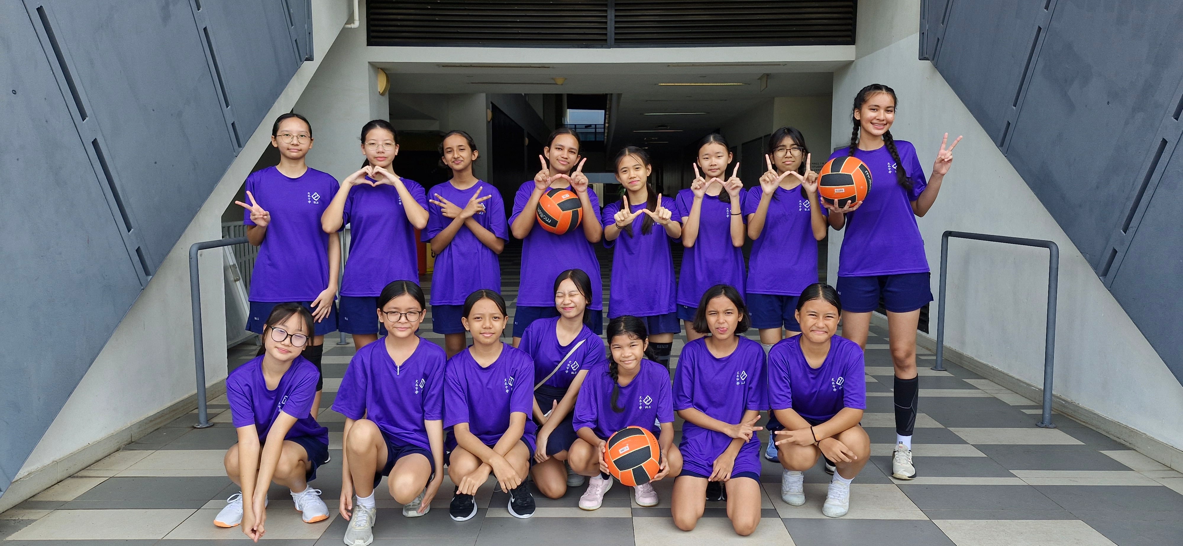 A group of girls in purple uniforms pose together holding orange volleyballs, making peace and heart signs.