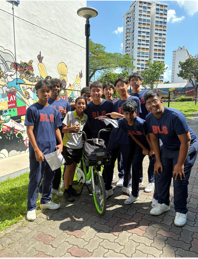 A group of boys in navy BLS uniforms pose together outdoors near a bicycle, with a colorful mural and tall buildings behind them.