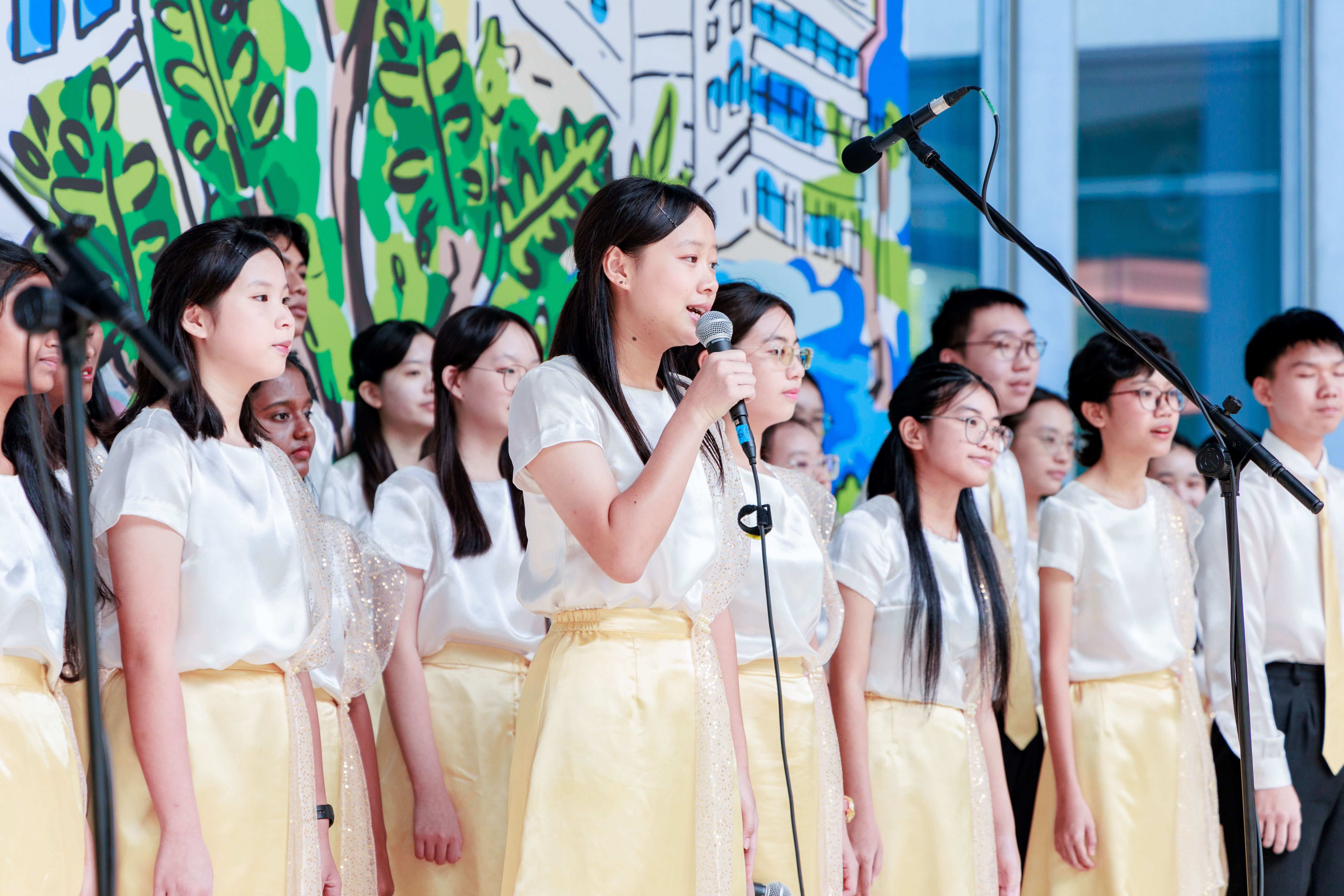 A youth choir in white tops and gold skirts performs on stage, with one girl singing into a handheld microphone.