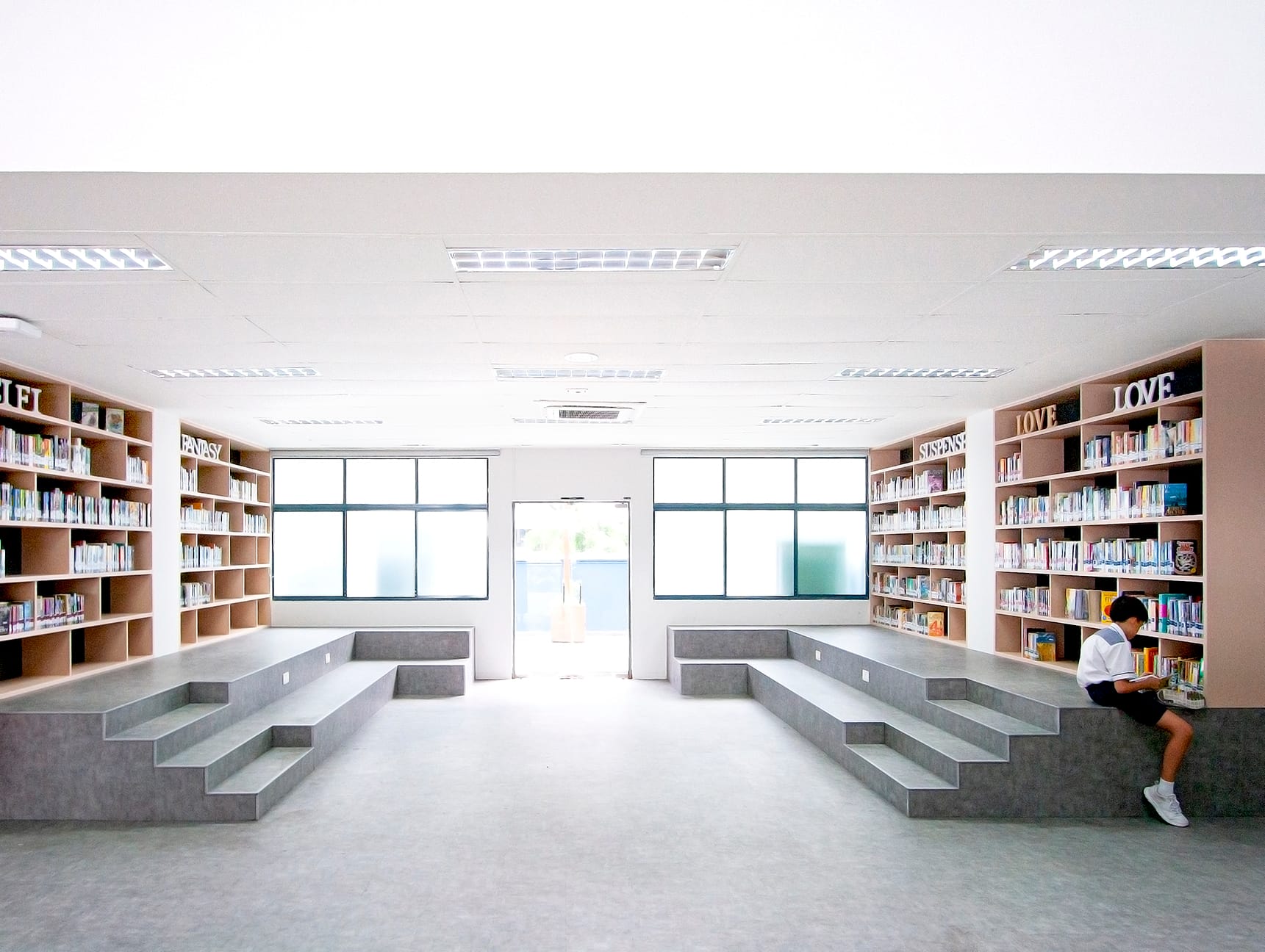 A bright school library with wood bookshelves, stepped seating platforms, and a child reading on the right side.