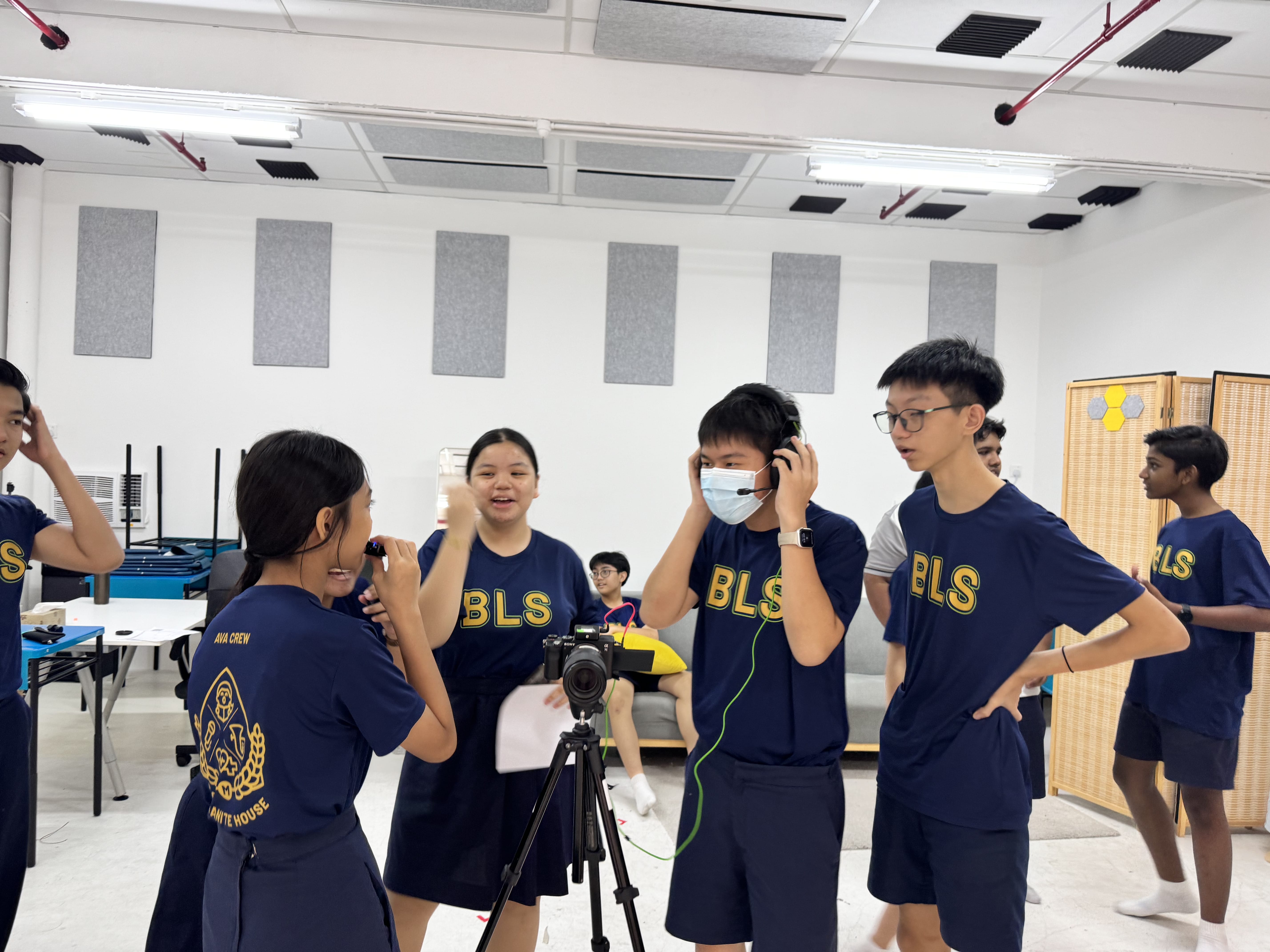 Students in navy BLS shirts gather around a camera on a tripod indoors, one wearing headphones and a face mask.