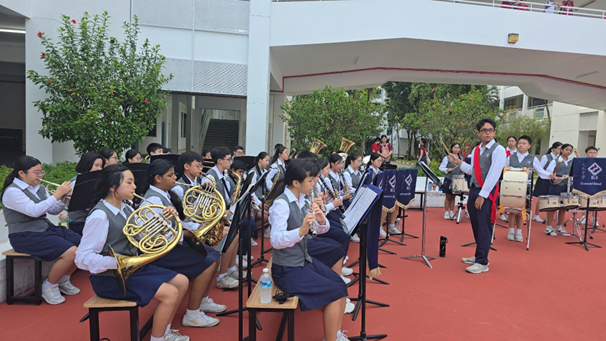 Students in uniforms perform as a school band outdoors, playing brass and wind instruments with a conductor standing nearby.