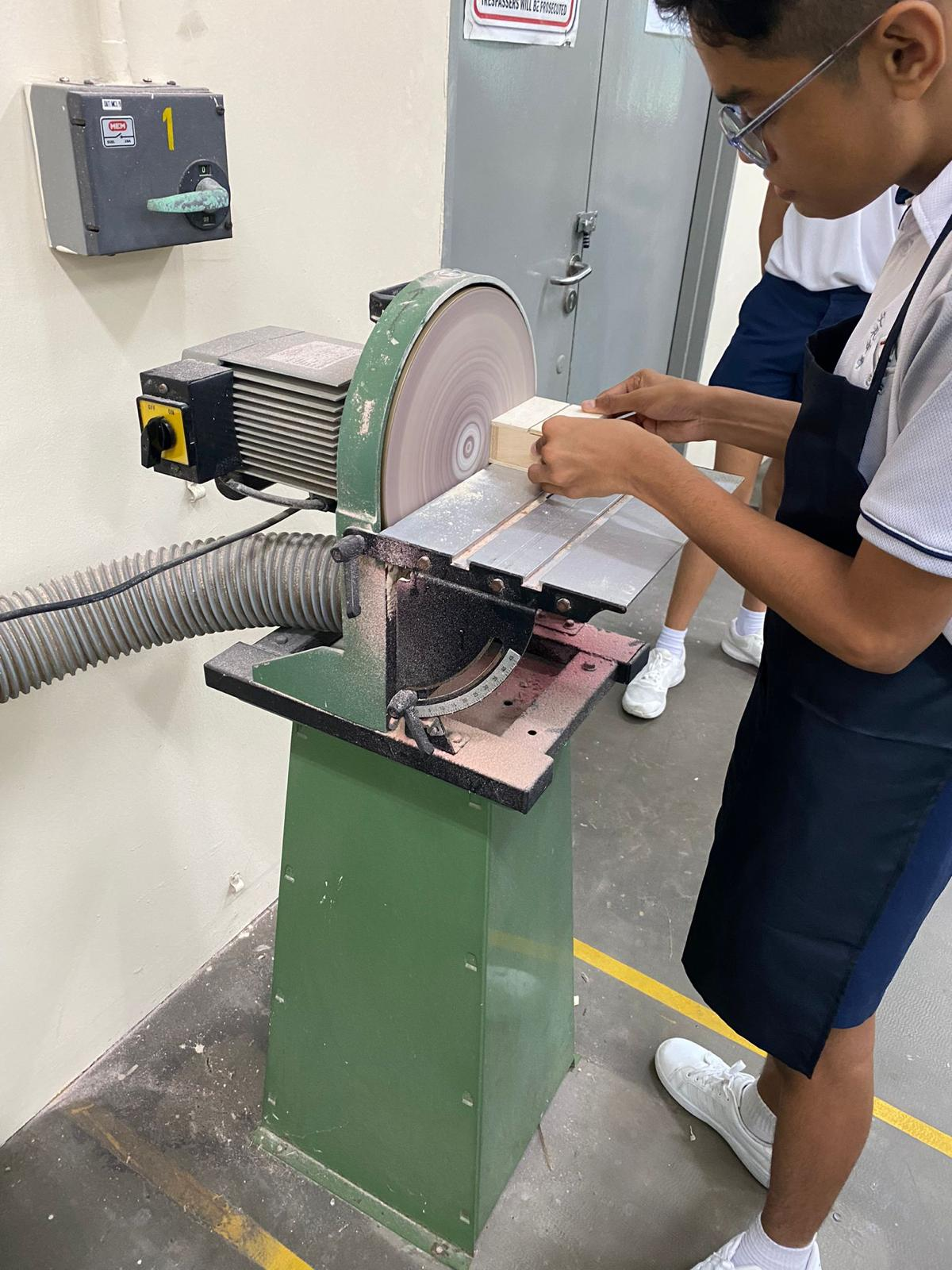 A young student in school uniform sanding a piece of wood on a green disc sander in a workshop.