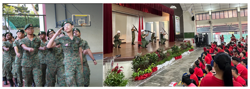 Students in camouflage uniforms march outdoors while soldiers perform a rifle drill on a stage before a seated audience.