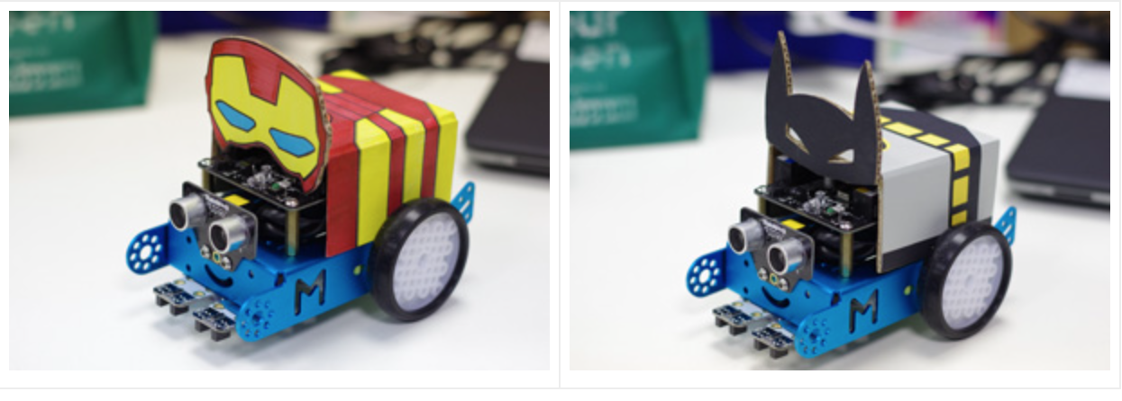 Two small wheeled robots decorated with cardboard Iron Man and Batman masks on a white surface.
