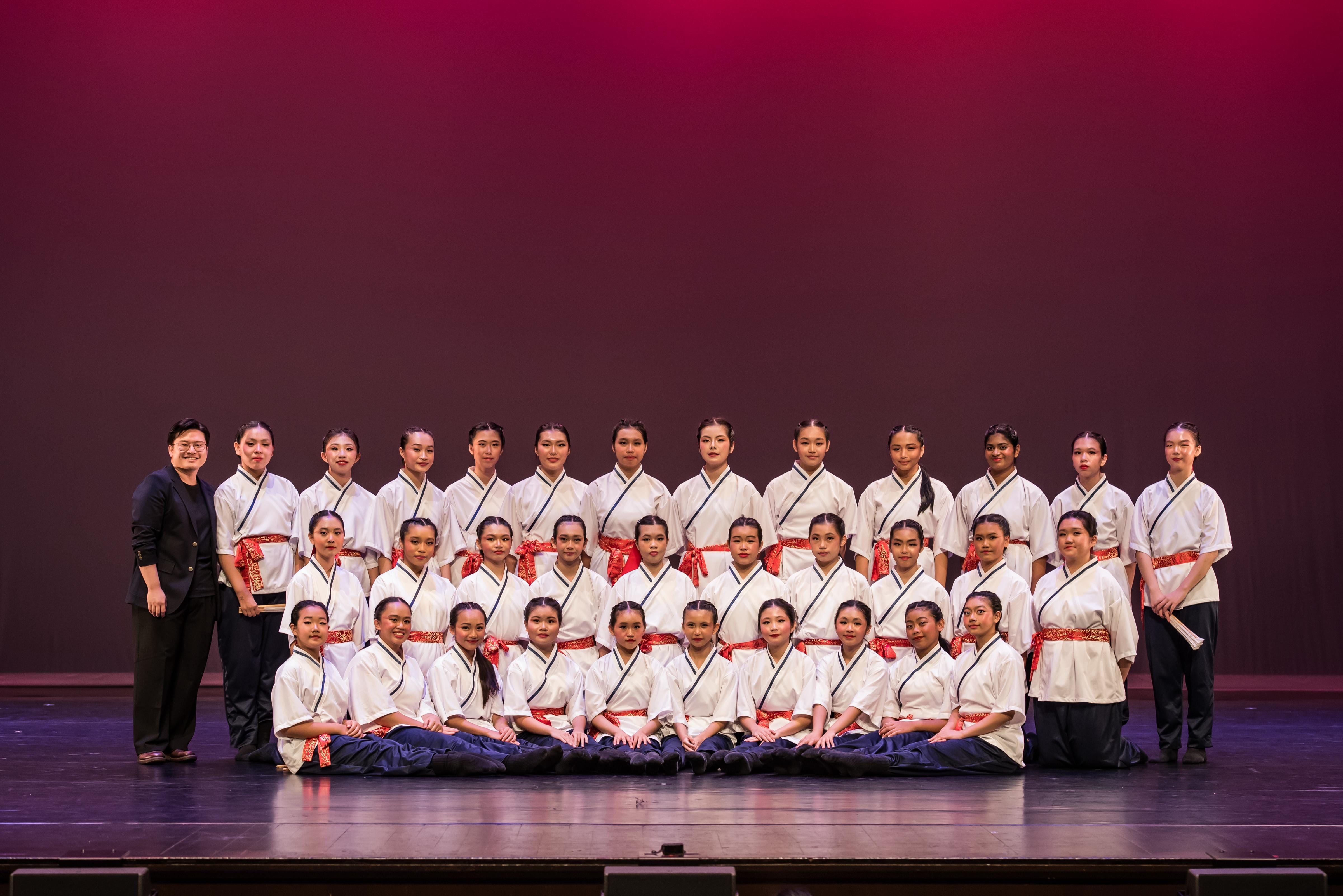 A large group of performers in white robes with red sashes pose in three rows on a stage with red lighting.