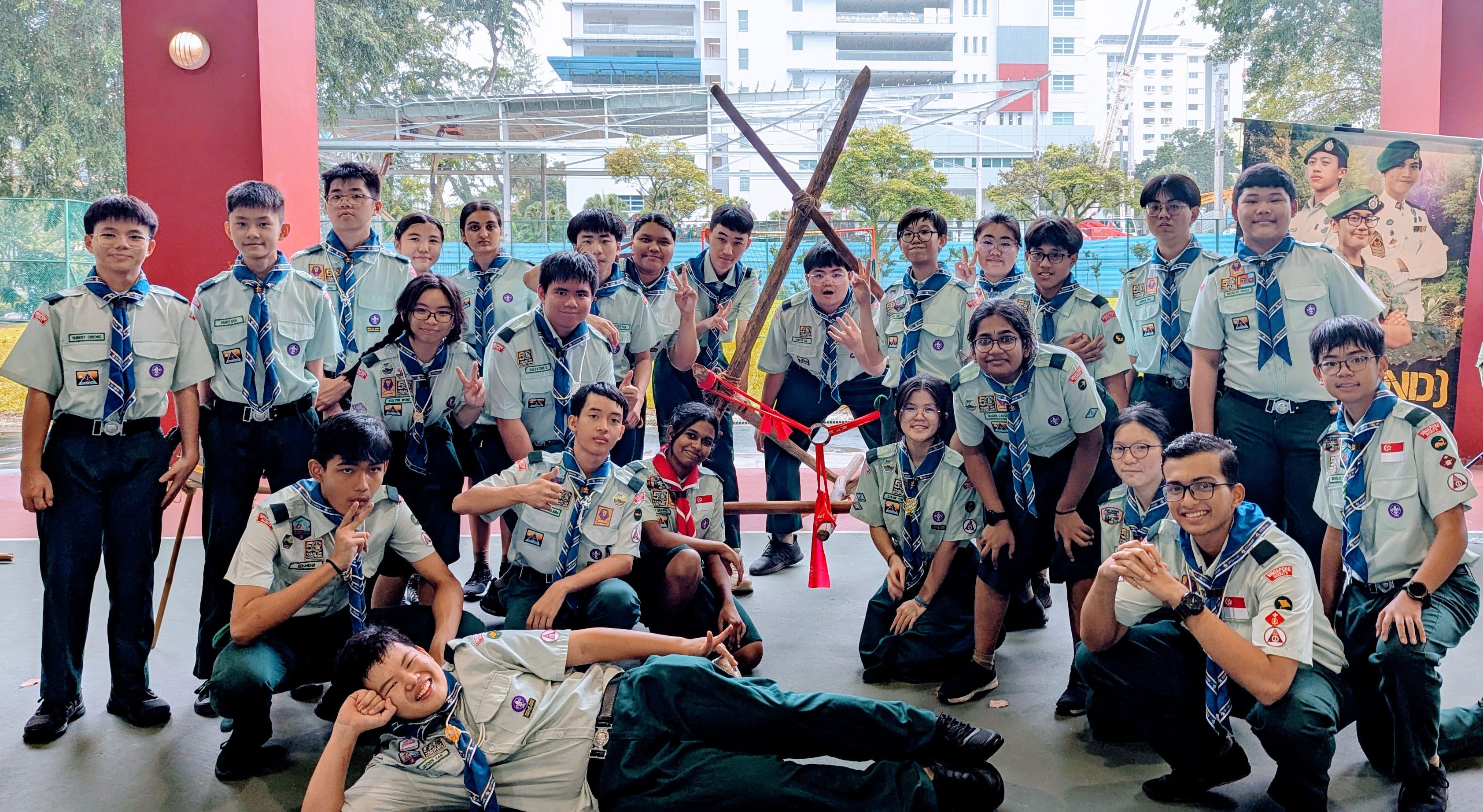 A large group of young scouts in uniform posing outdoors with a wooden pioneering structure tied with red lashings.