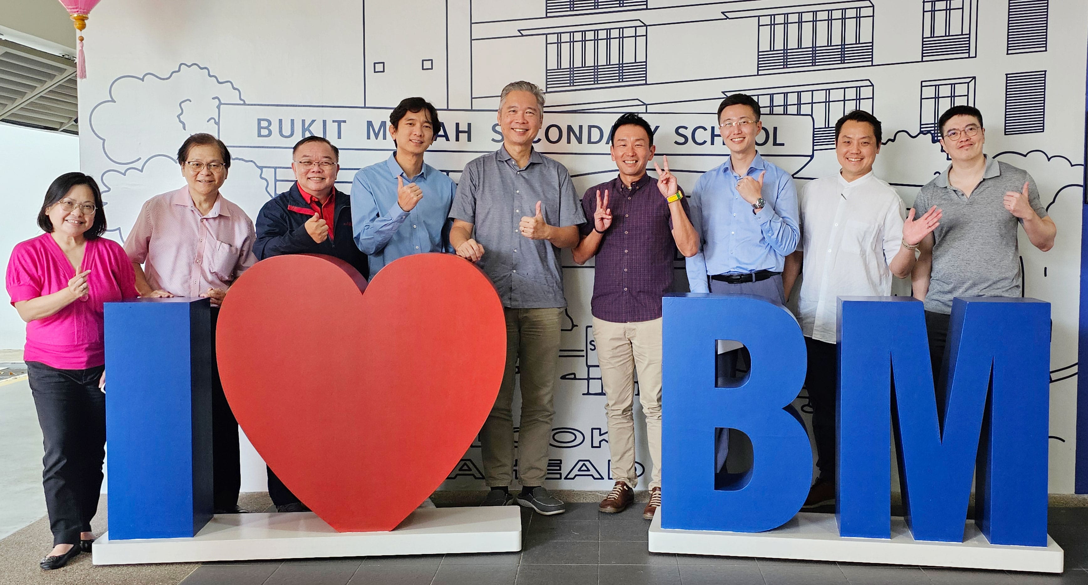 Nine people posing behind large blue letters spelling I heart BM at Bukit Merah Secondary School.