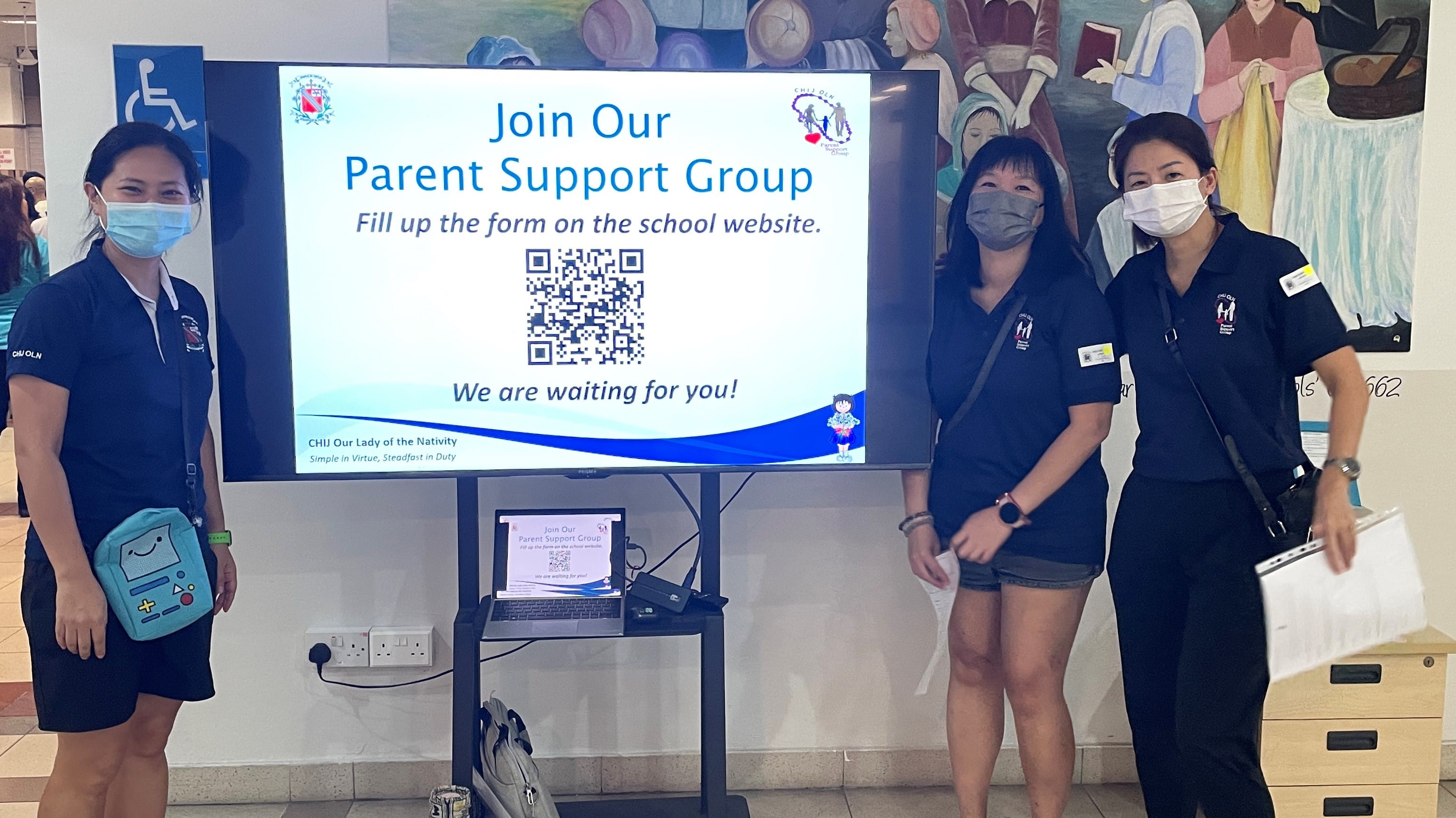 Three masked women in navy uniforms stand beside a screen displaying a Join Our Parent Support Group sign with a QR code.