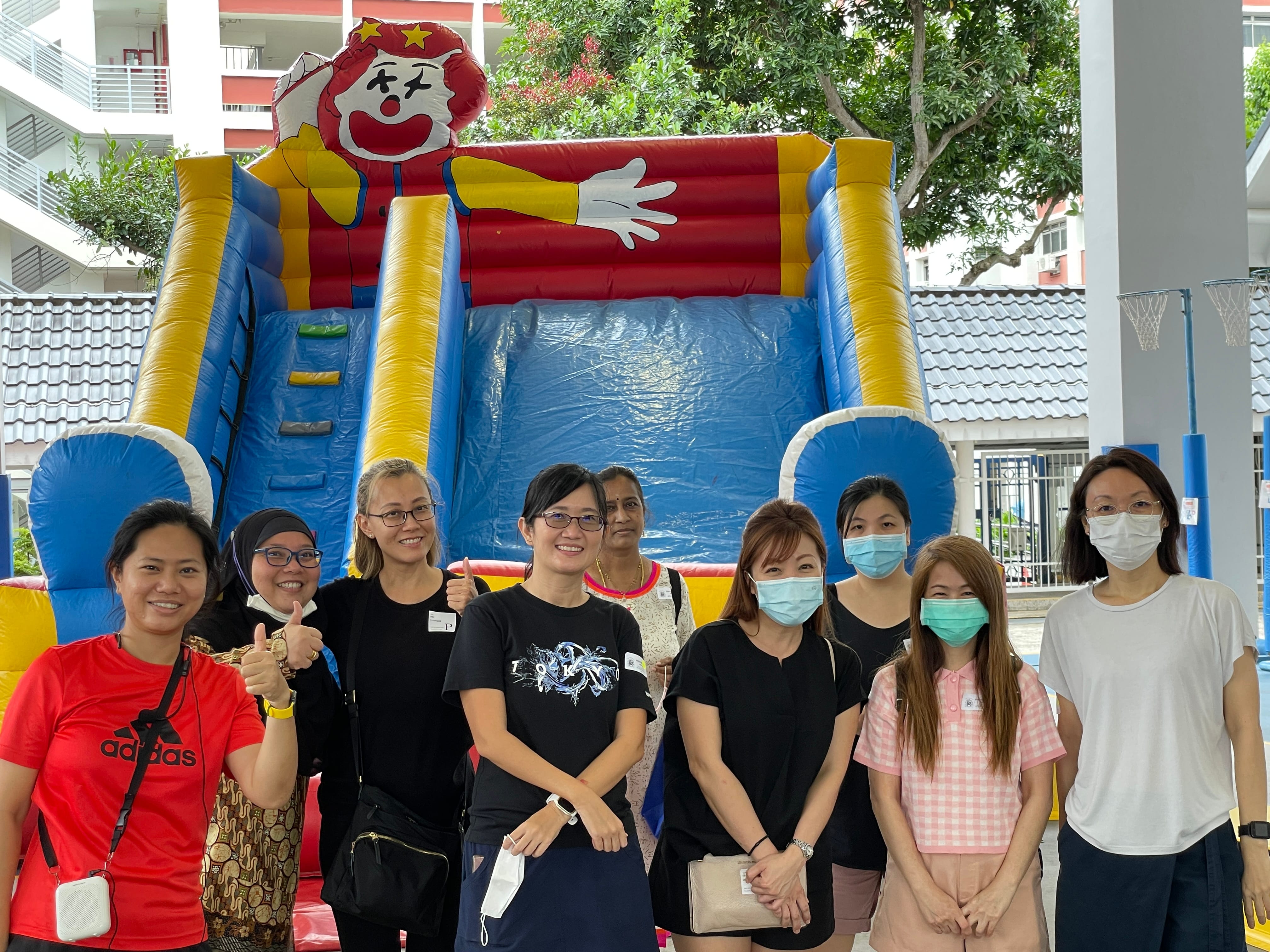 Nine women posing in front of a colorful inflatable clown slide at an outdoor event.