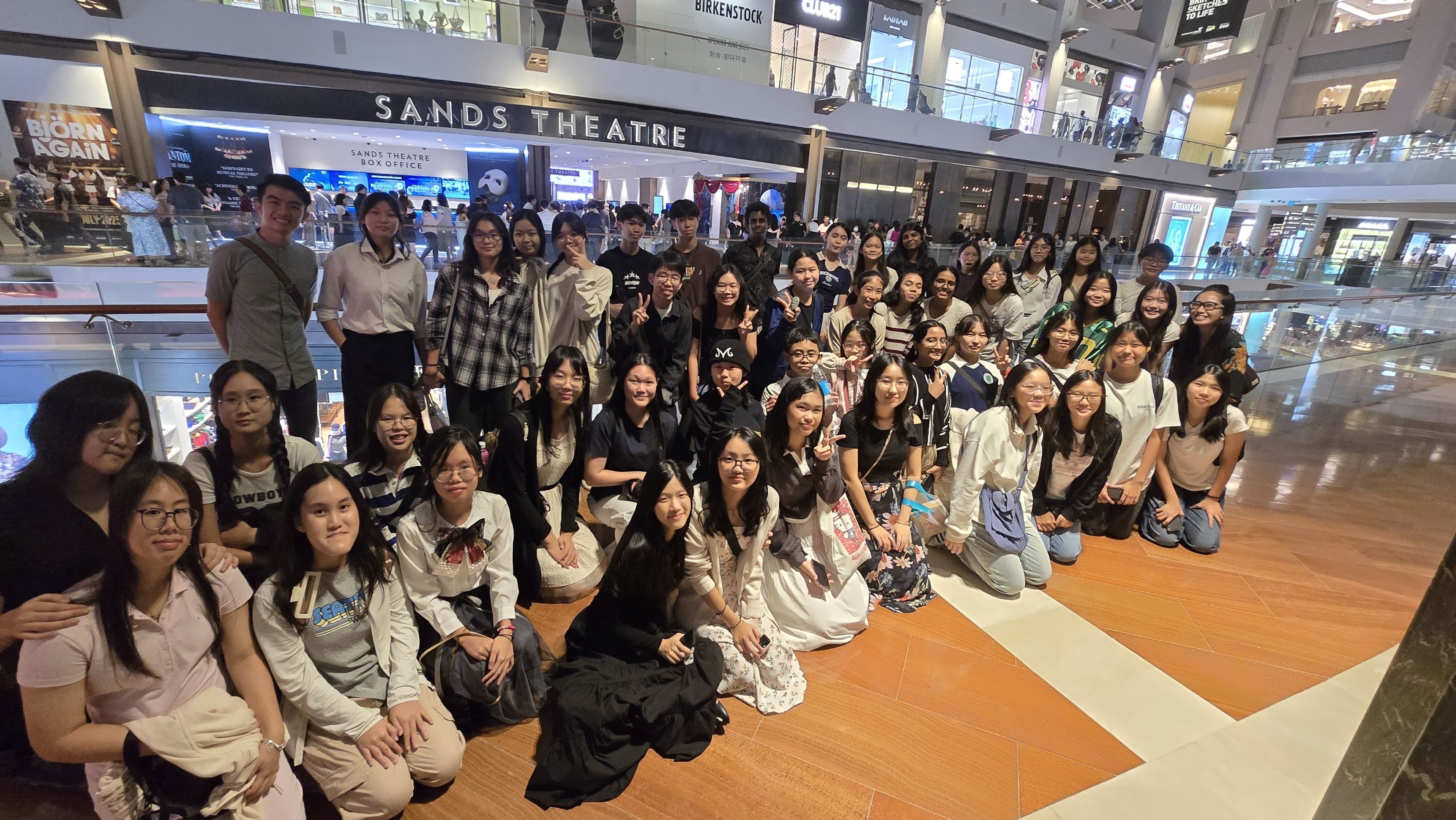 Large group of young people posing together inside a shopping mall in front of Sands Theatre signage.