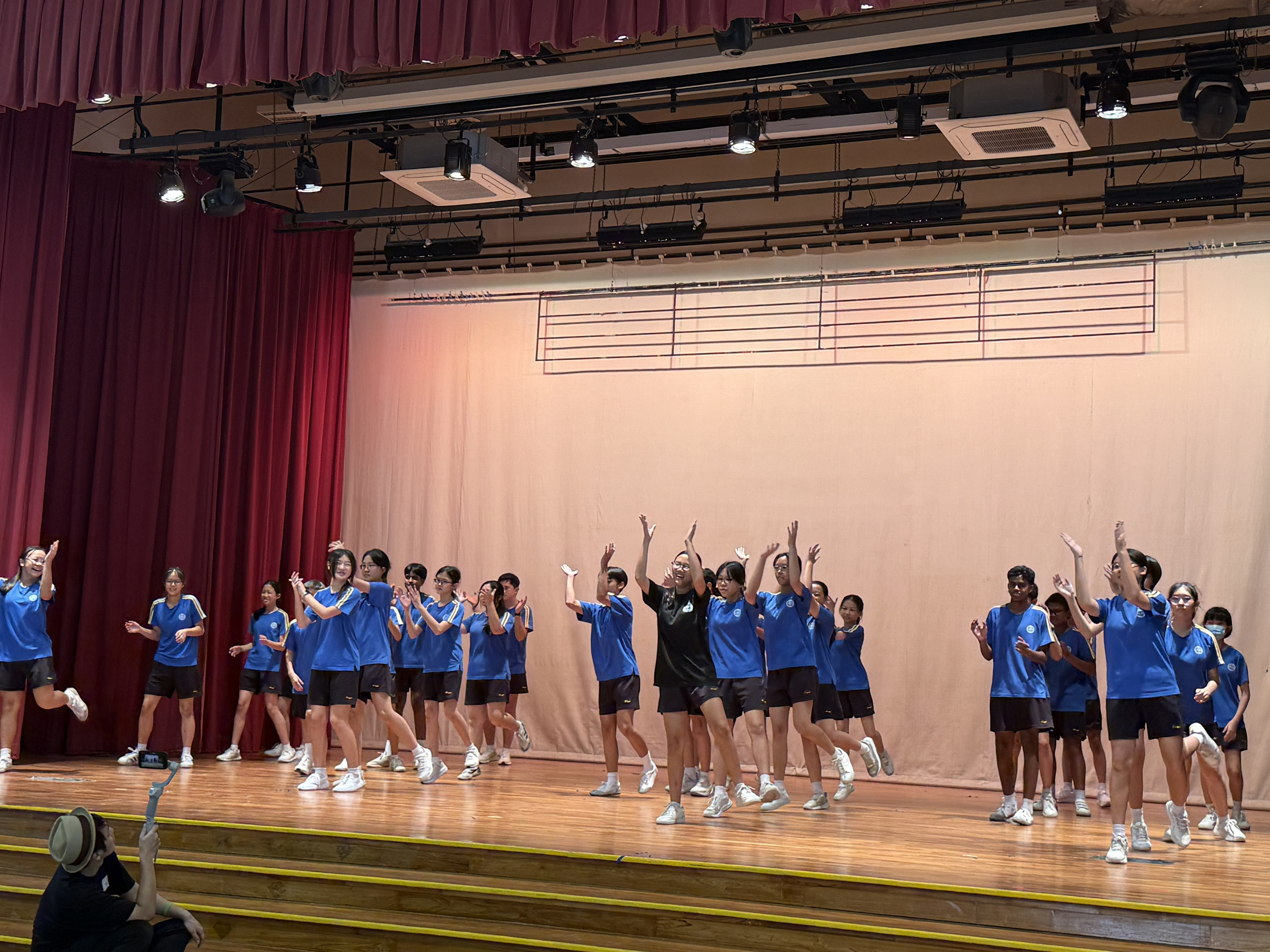 A large group of students in blue uniforms dancing on a wooden stage with red curtains and stage lights above.