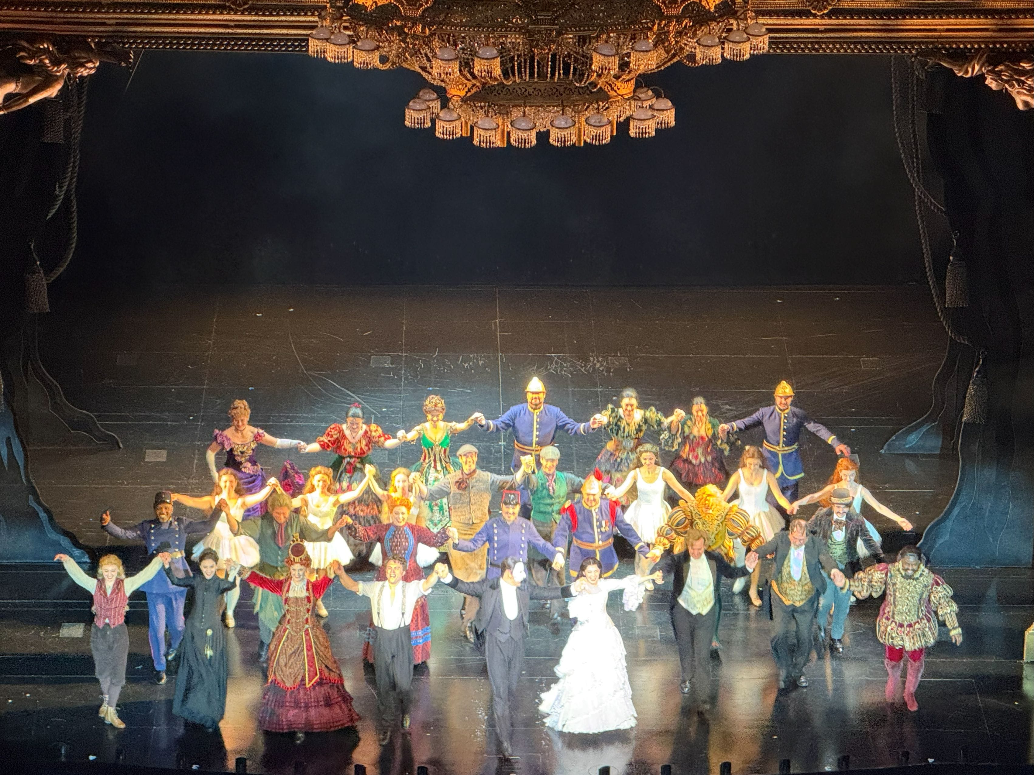 A large cast in colorful period costumes holds hands and takes a curtain call bow on an ornate theater stage.