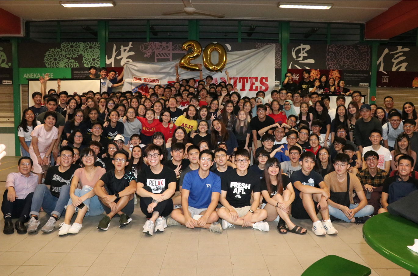 Large group of students posing indoors under a gold number 20 balloon with a banner behind them.
