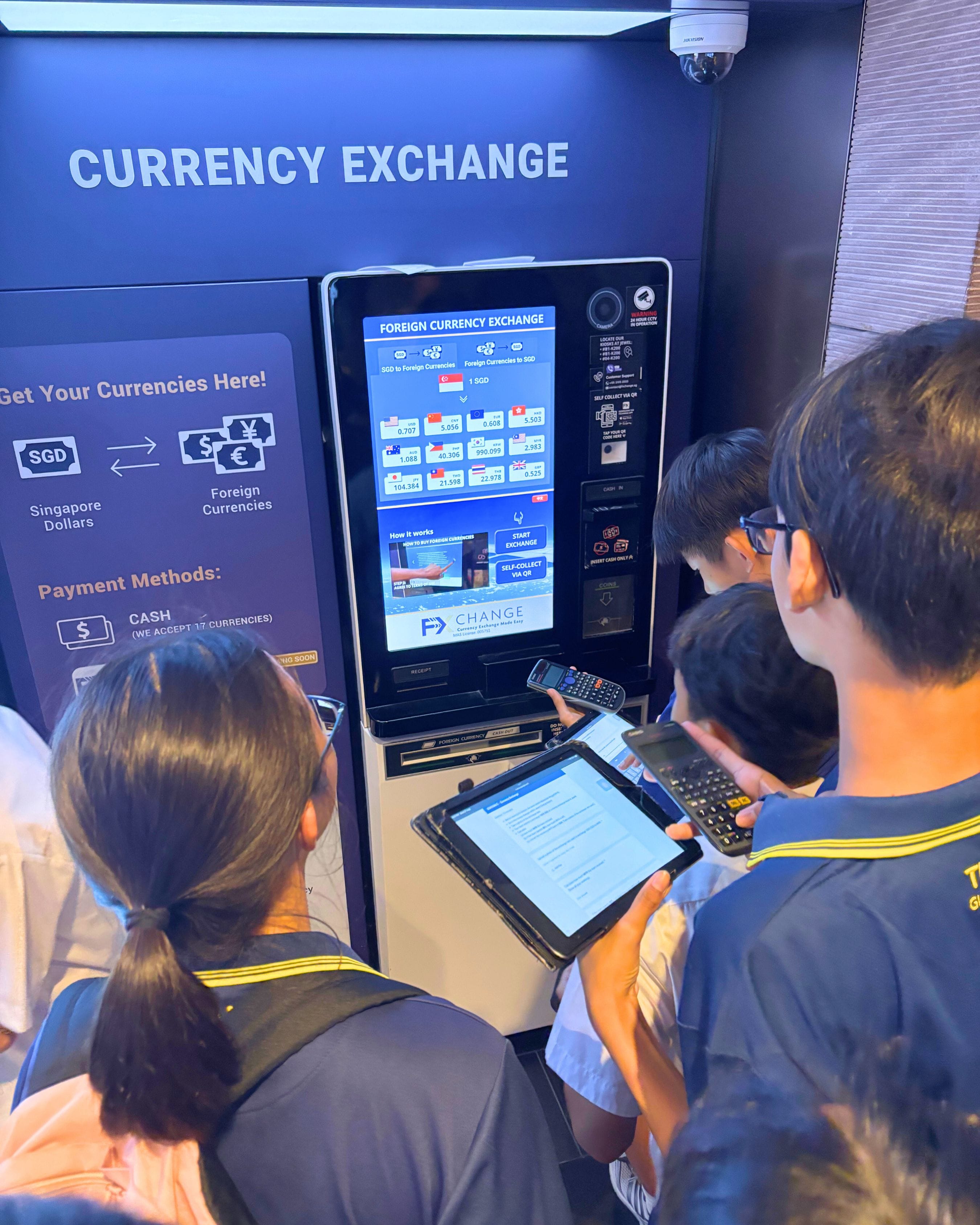 Three students in uniforms look at a blue foreign currency exchange kiosk, one holding a tablet.