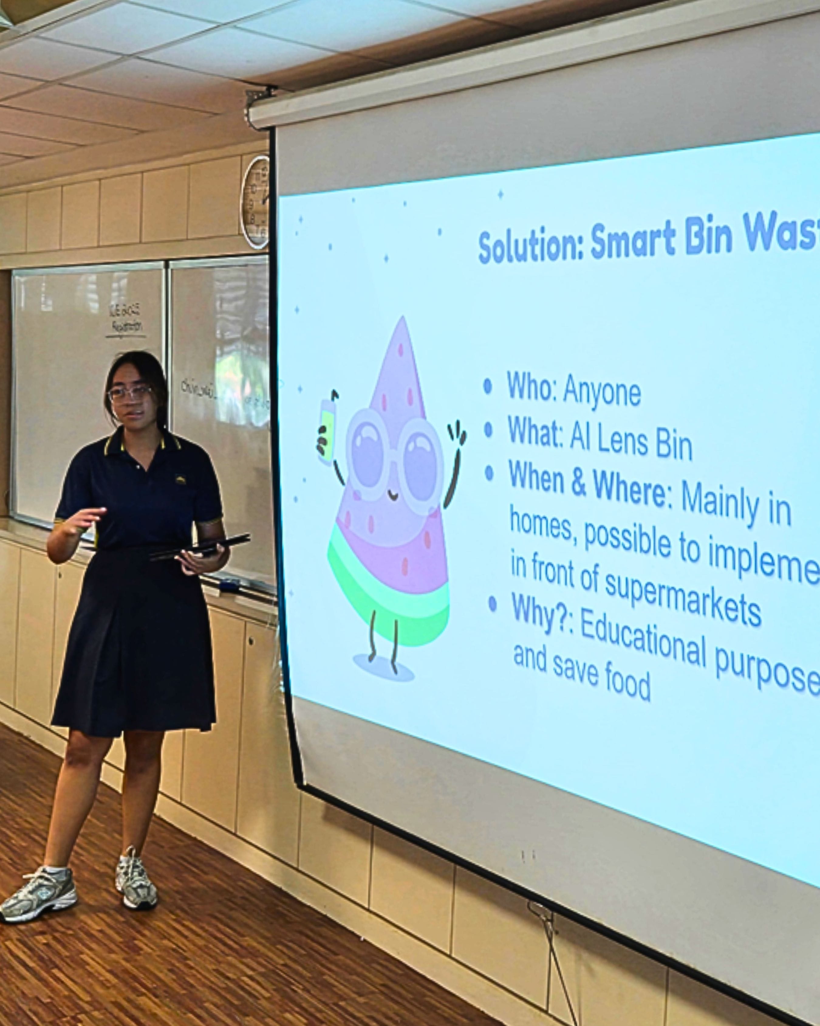 A student in a navy uniform presents beside a projected slide titled Solution: Smart Bin Waste with bullet points.