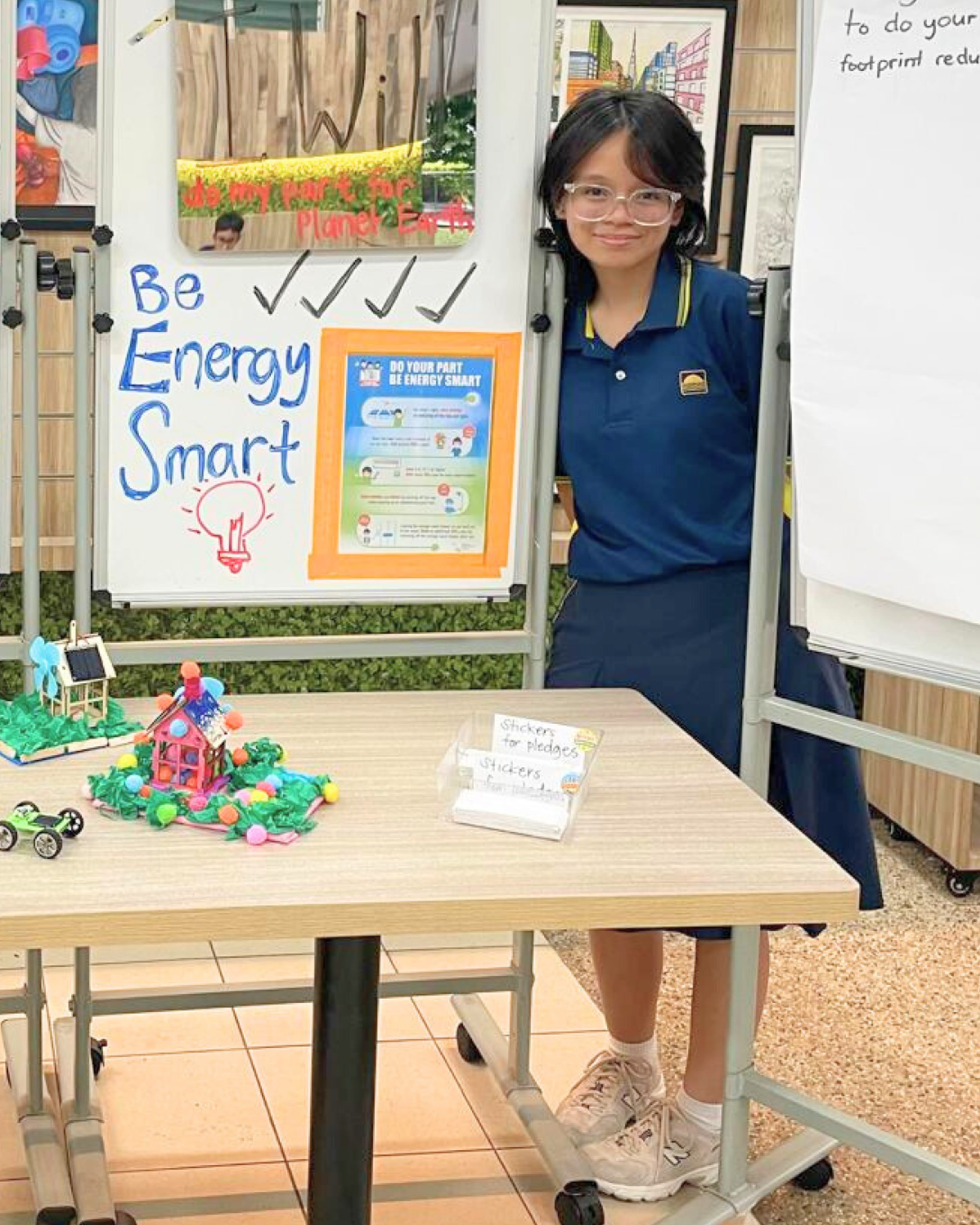 Student stands by a display board reading Be Energy Smart, with a table of models and pledge stickers.