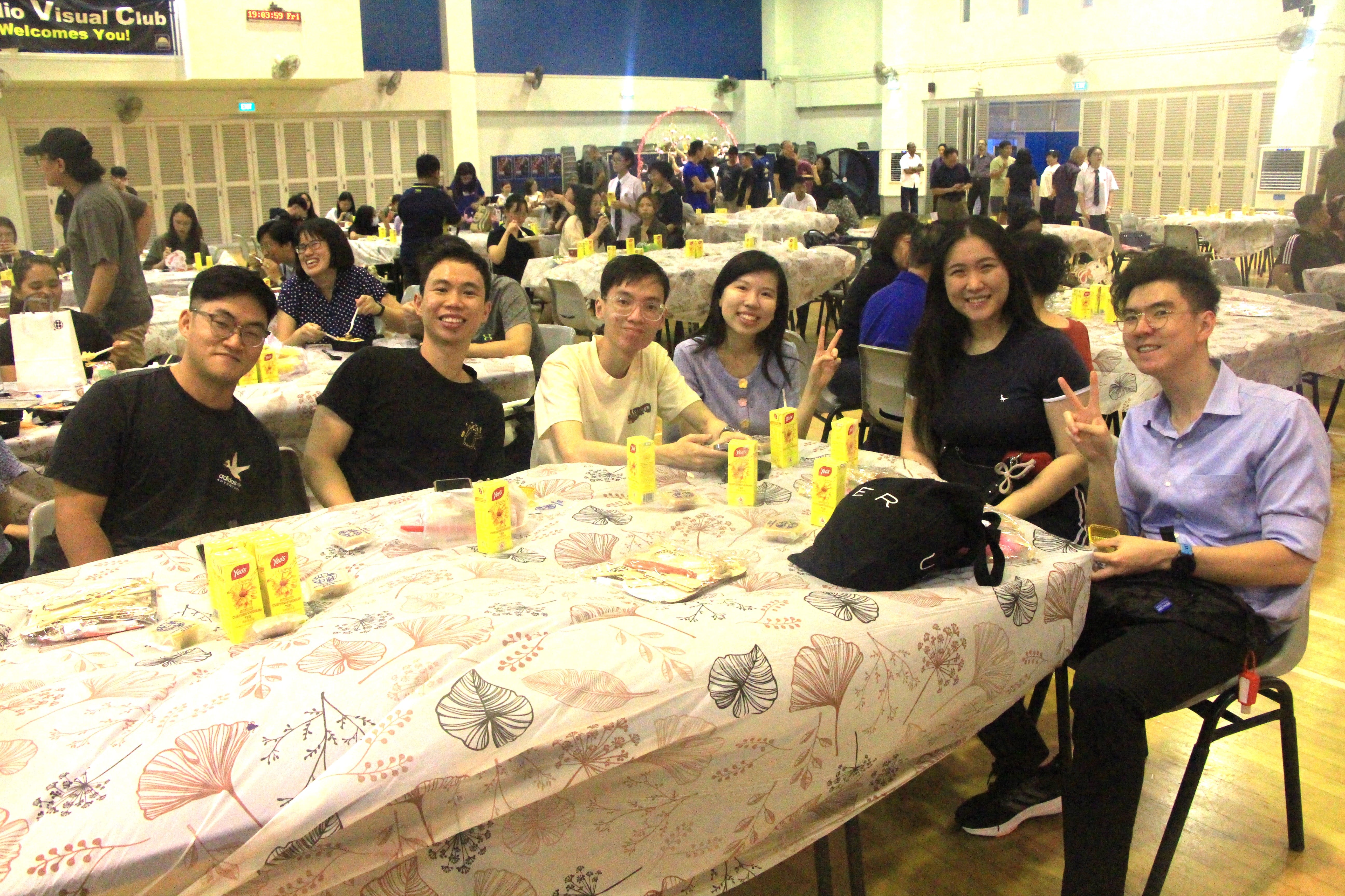 Six people smile at a banquet table with yellow drink cartons, in a crowded hall with many tables.