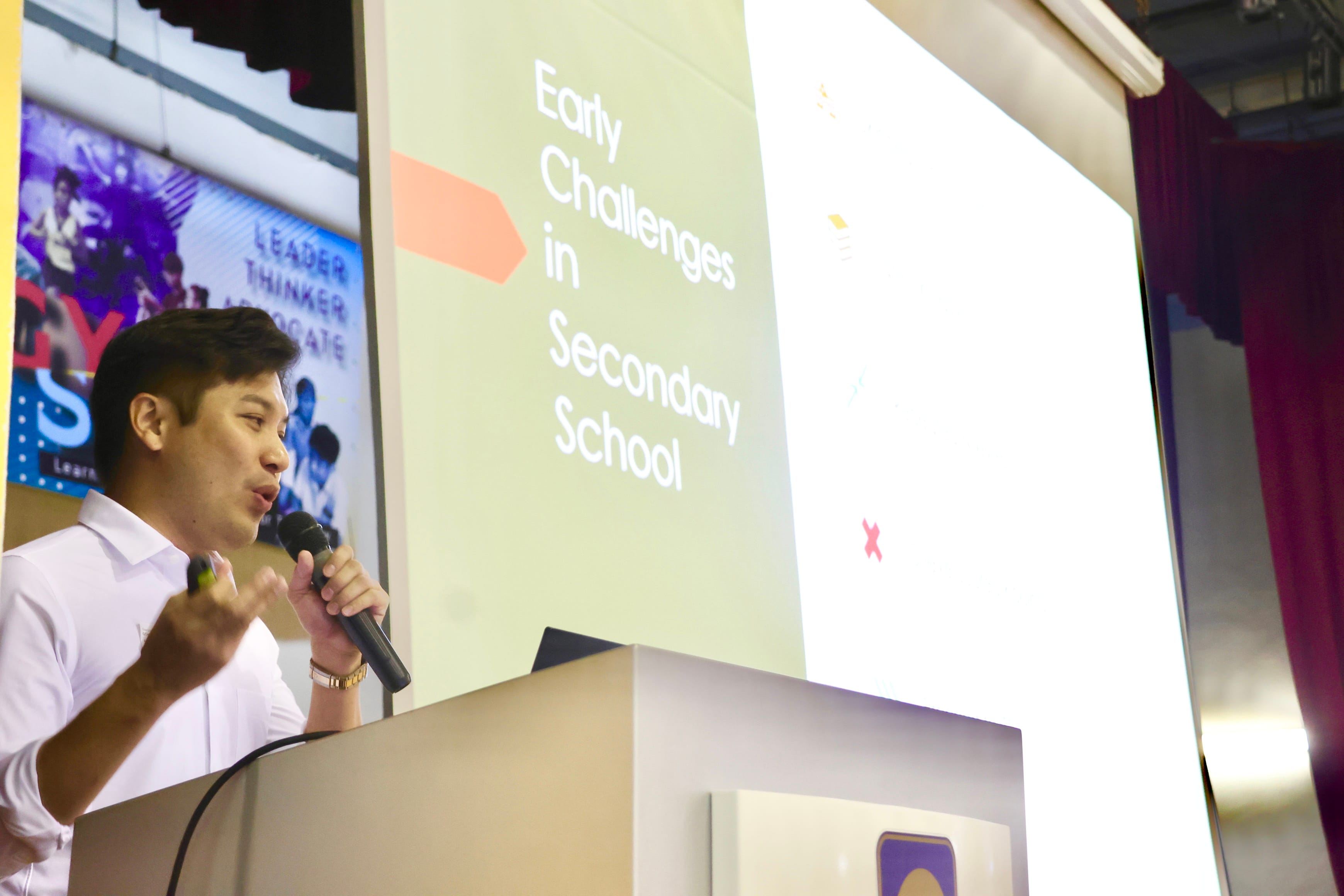 A man in a white shirt speaks at a podium with a microphone beside a slide reading "Early Challenges in Secondary School."