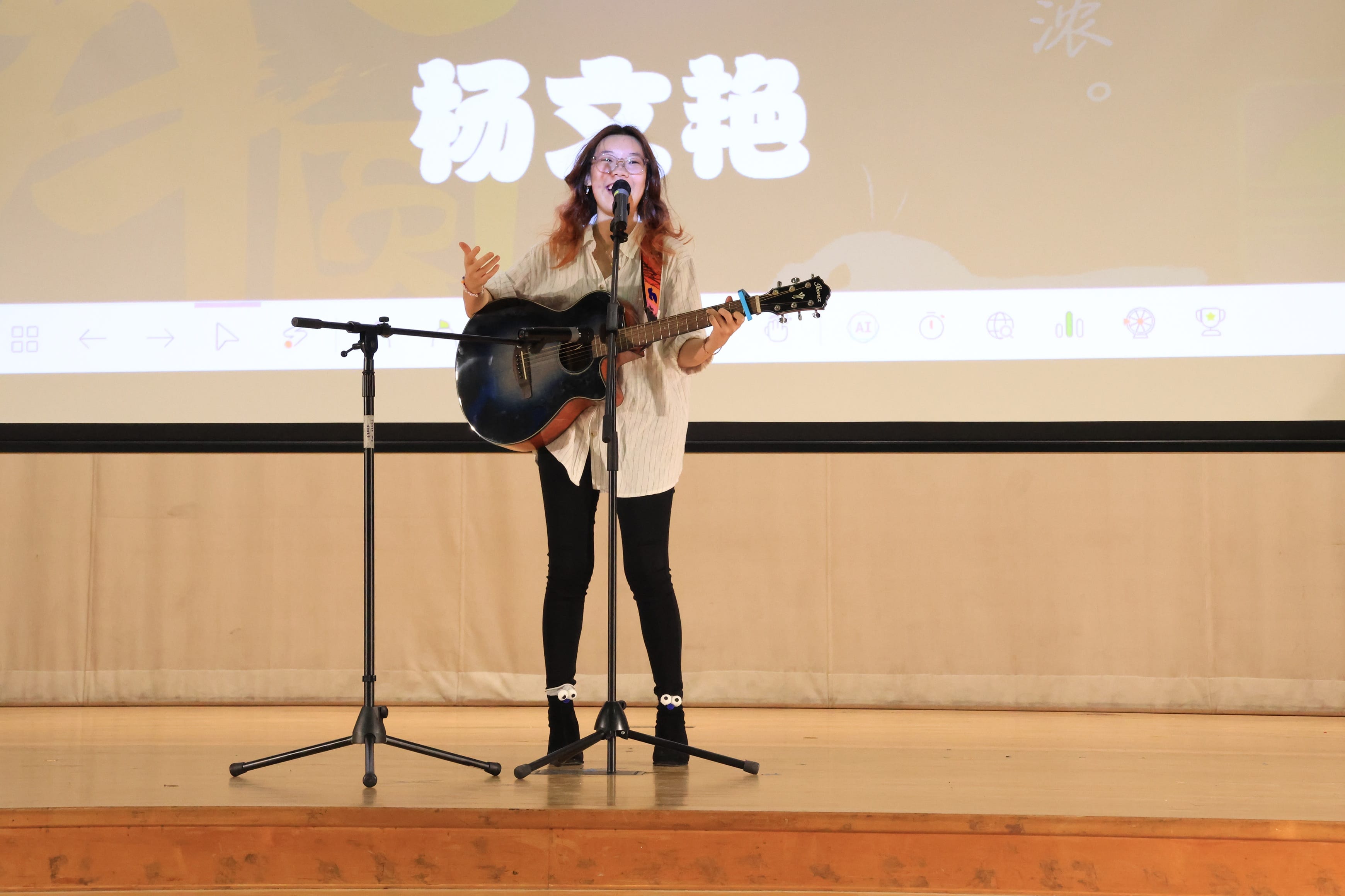 A young woman with glasses performs on stage, singing into a mic while holding a black acoustic guitar.