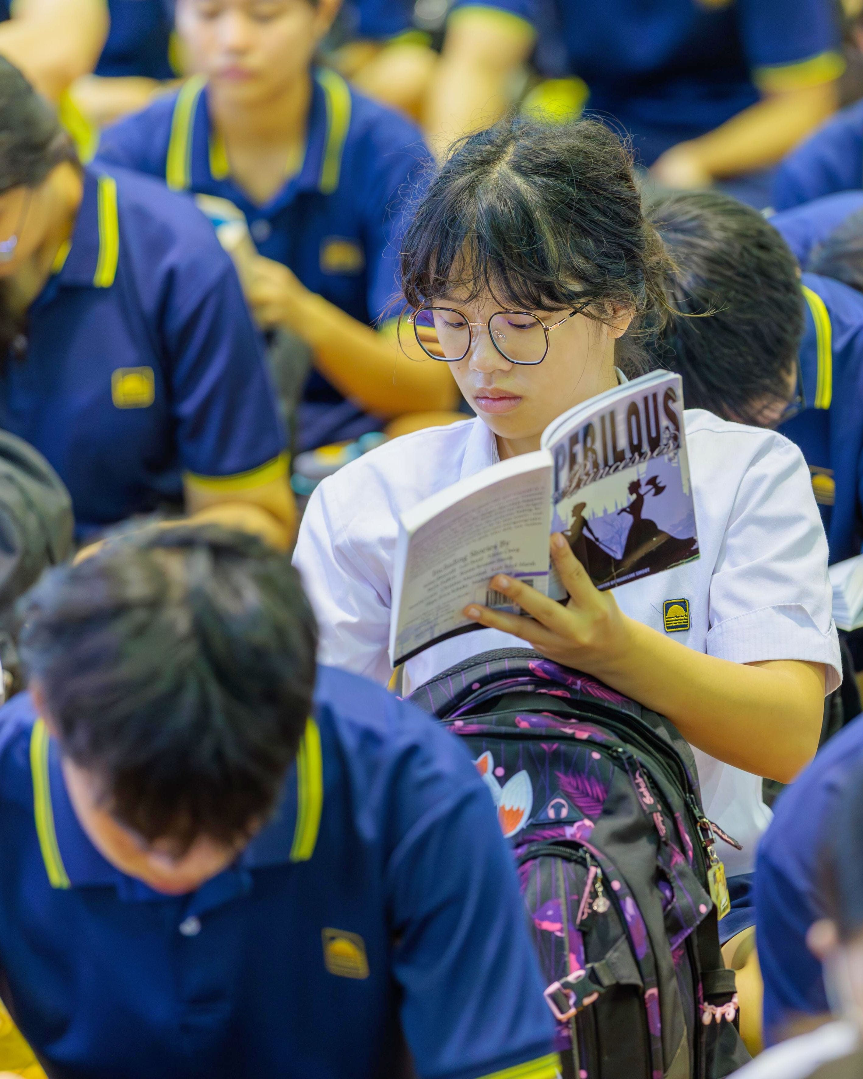 A teenage girl with glasses in a white uniform reads a book titled Perilous, surrounded by students in blue uniforms.