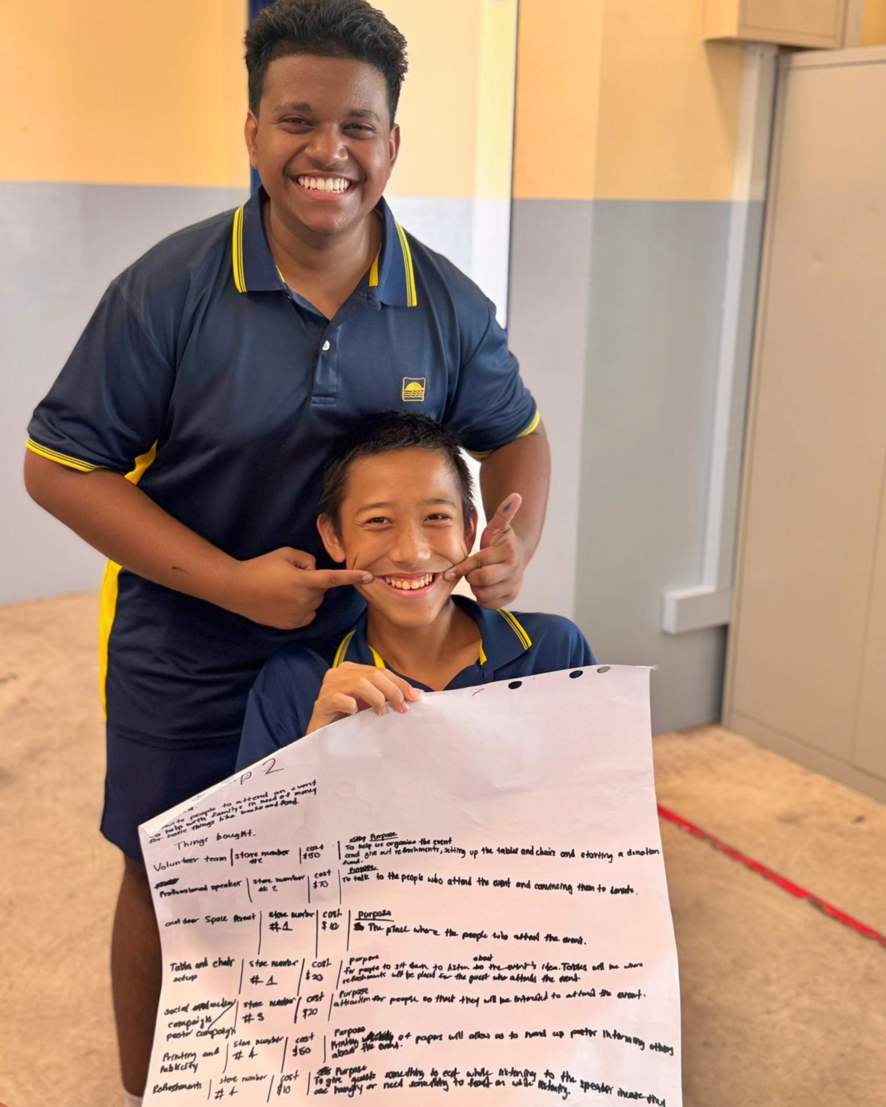 Two smiling students in navy and yellow uniforms hold a handwritten planning sheet with tables and notes.