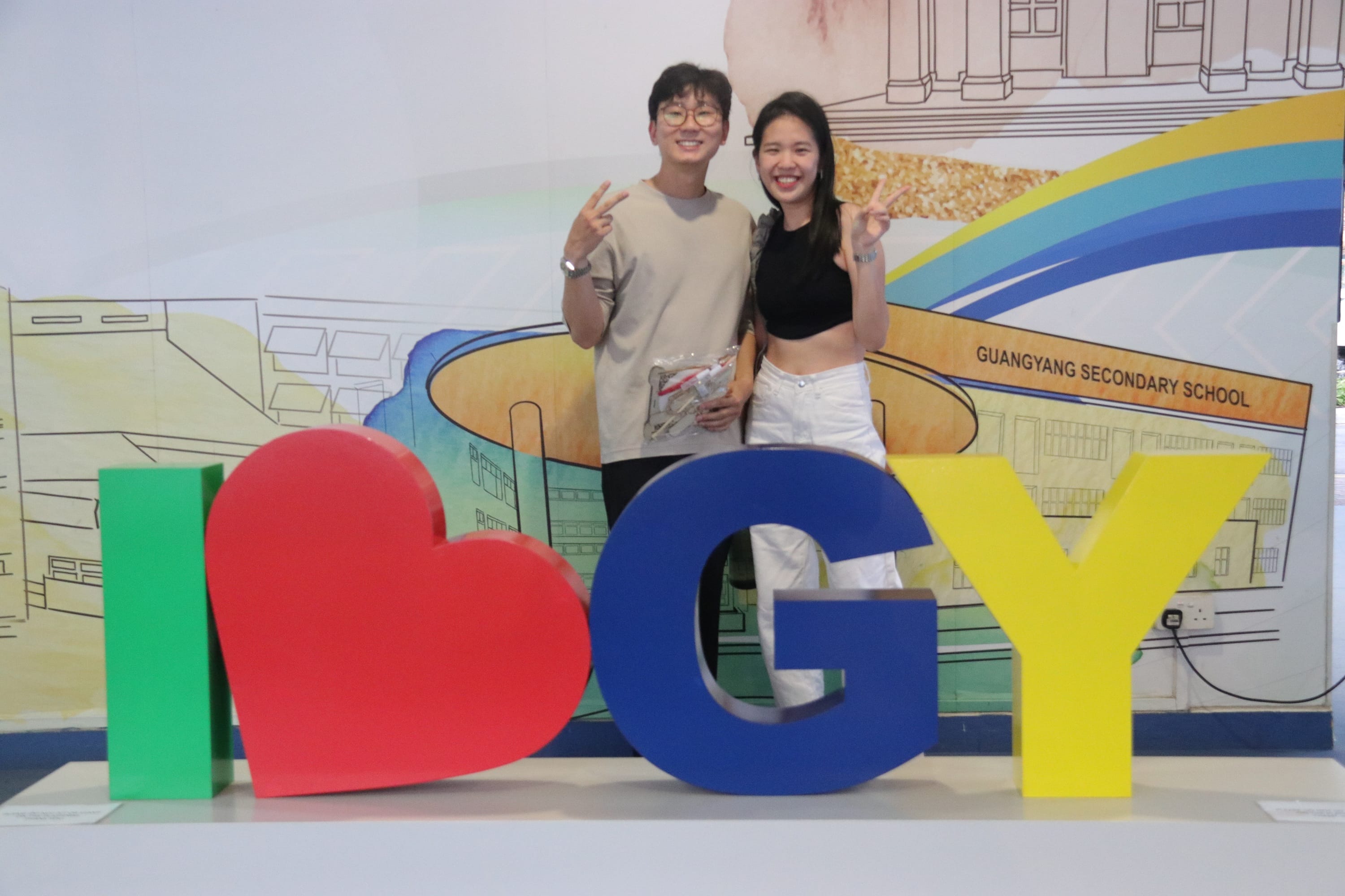 Two smiling people pose with peace signs behind colorful I Love GY letters at Guangyang Secondary School.