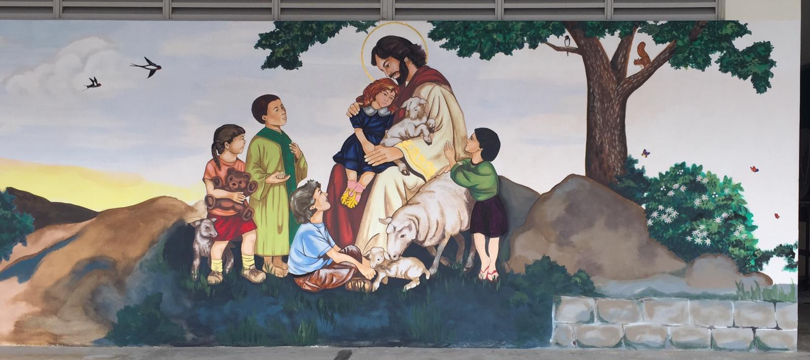 Outdoor mural of Jesus with a halo embracing children and lambs, surrounded by trees, rocks, and butterflies.