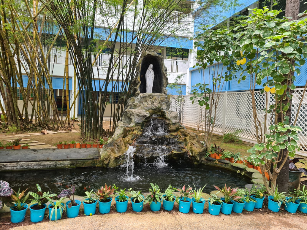 A garden grotto with a white religious statue, waterfall into a pond, blue pots, and bamboo trees near a blue building.