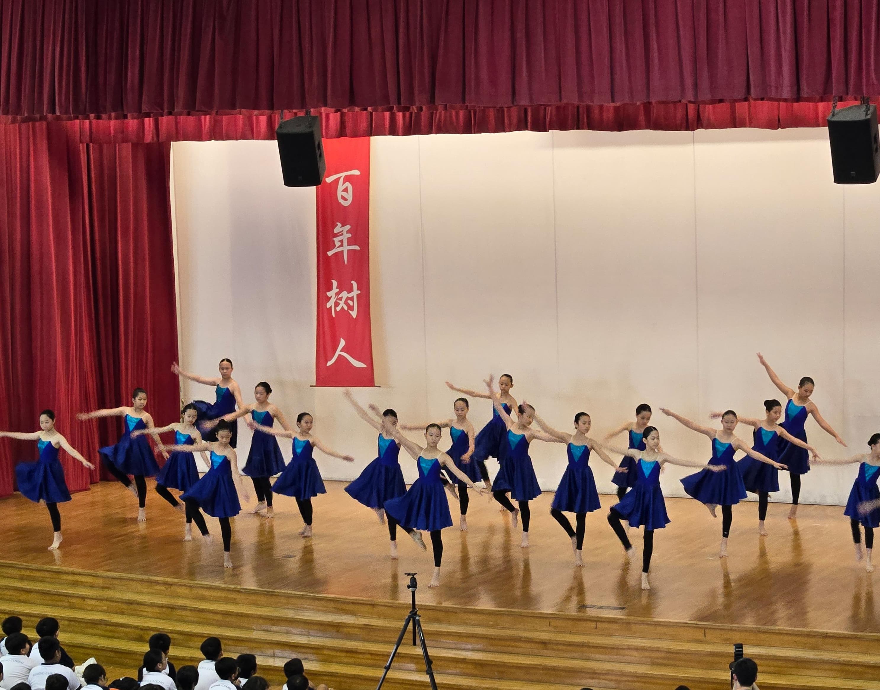 Young girls in blue dresses perform a synchronized dance on a wooden stage with red curtains and a Chinese banner.
