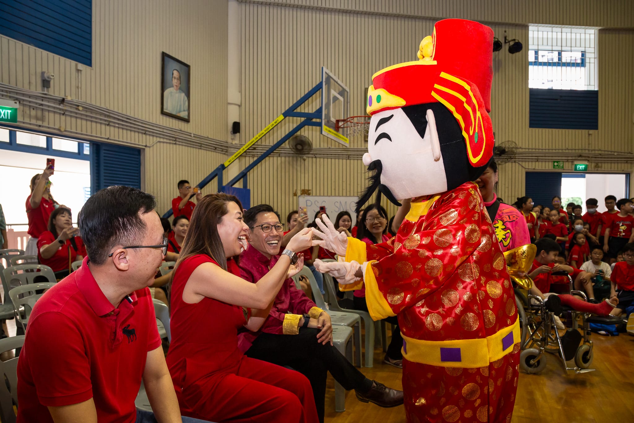 A red-costumed Chinese deity mascot interacts with laughing seated adults in red shirts inside a school hall.