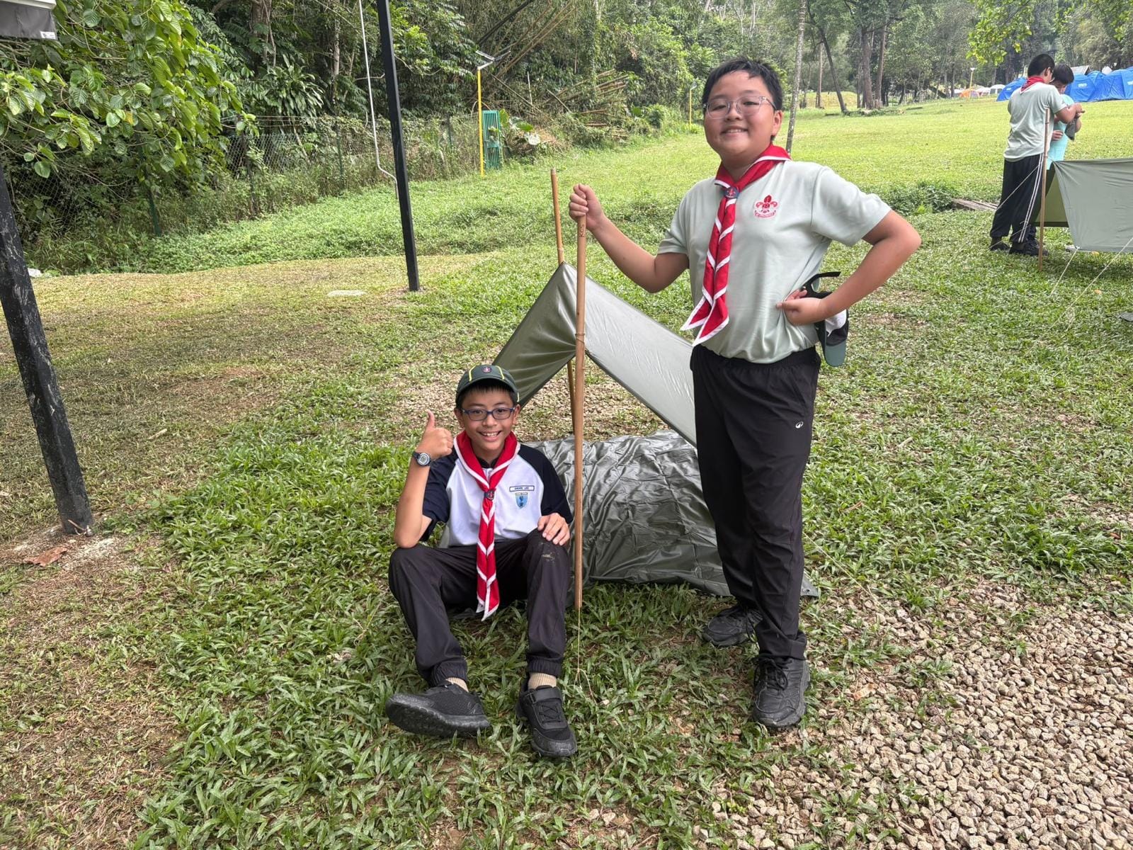 Two young scouts in uniform pose beside a small green tent on grass at an outdoor campsite.