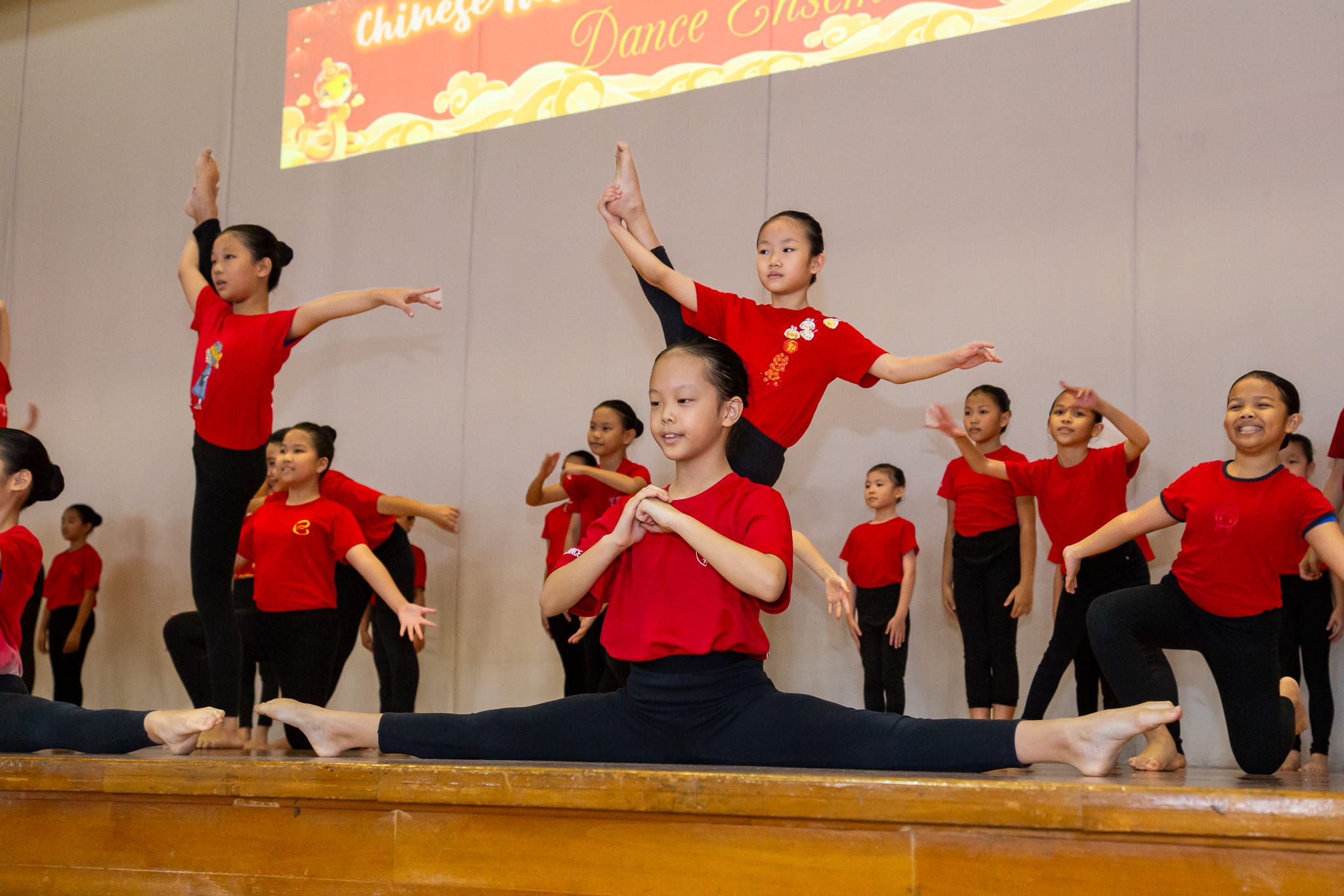 Children in red shirts perform a Chinese dance ensemble on stage, one doing full splits while another balances above.