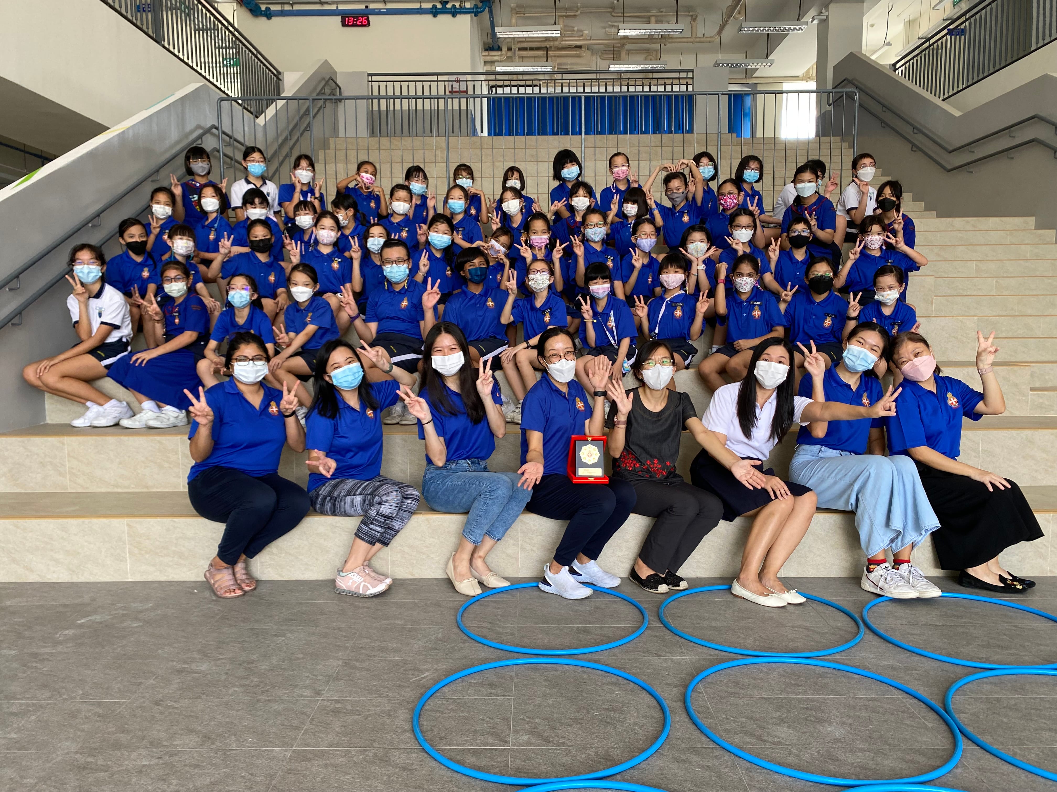 Group of masked students in blue uniforms on steps, many flashing peace signs; blue hoops on floor.