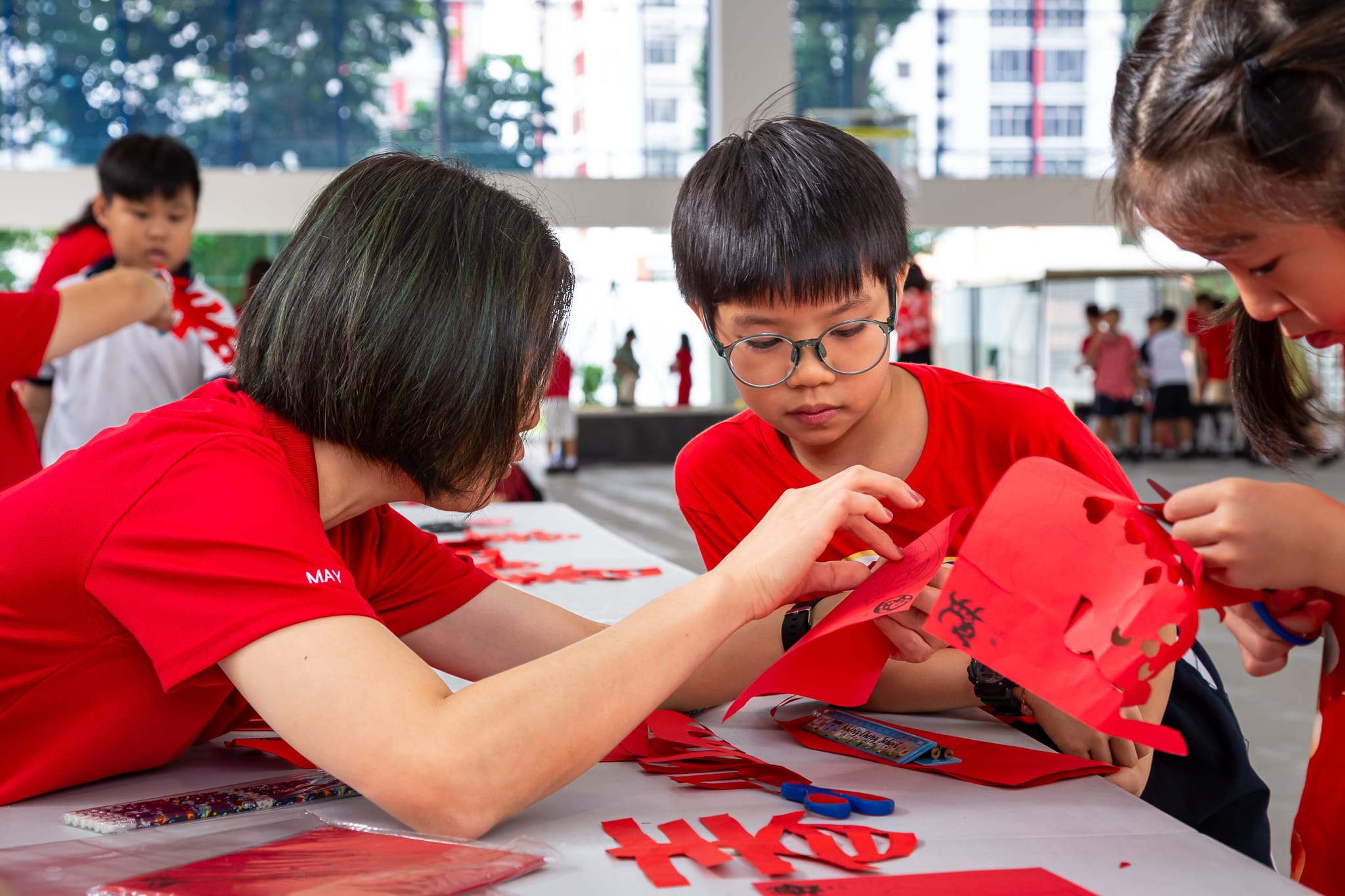 Children and an adult in red shirts work together on red paper crafts at a table outdoors.