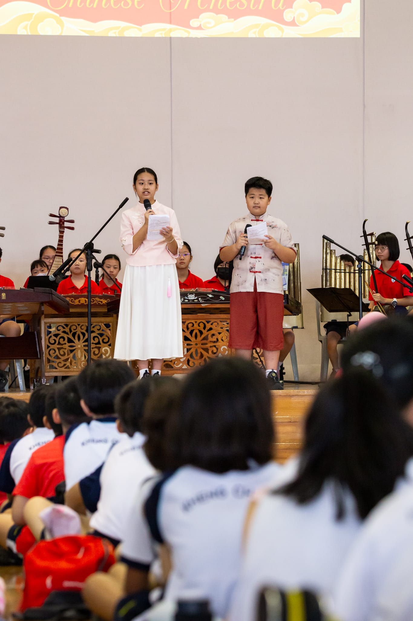 Two students hold microphones and papers on stage before a seated audience, with a Chinese orchestra performing behind them.