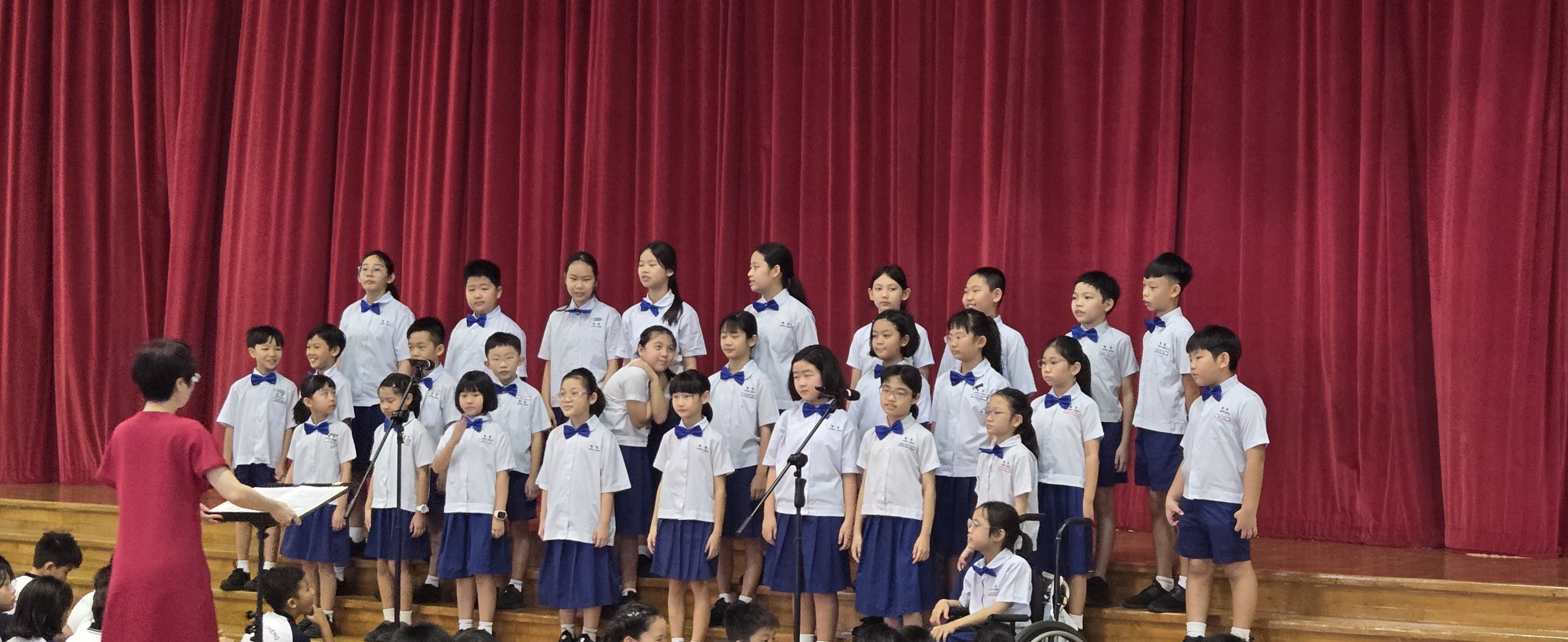 Children in white shirts and blue bow ties perform as a choir on stage before a red curtain, led by a conductor.