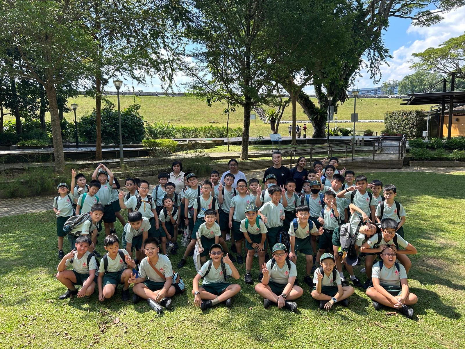 A large group of schoolboys in matching green uniforms pose with teachers on a grassy park lawn surrounded by trees.