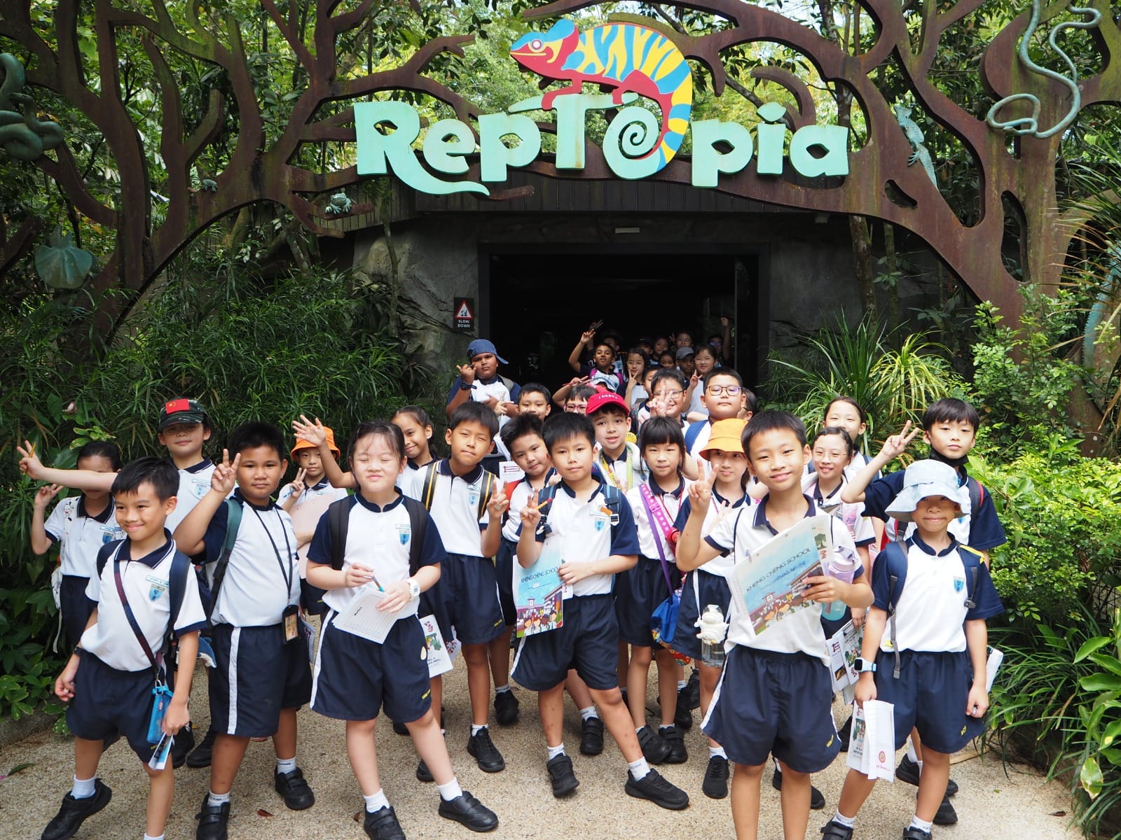 Group of young students in school uniforms posing with peace signs at the entrance of Reptopia.